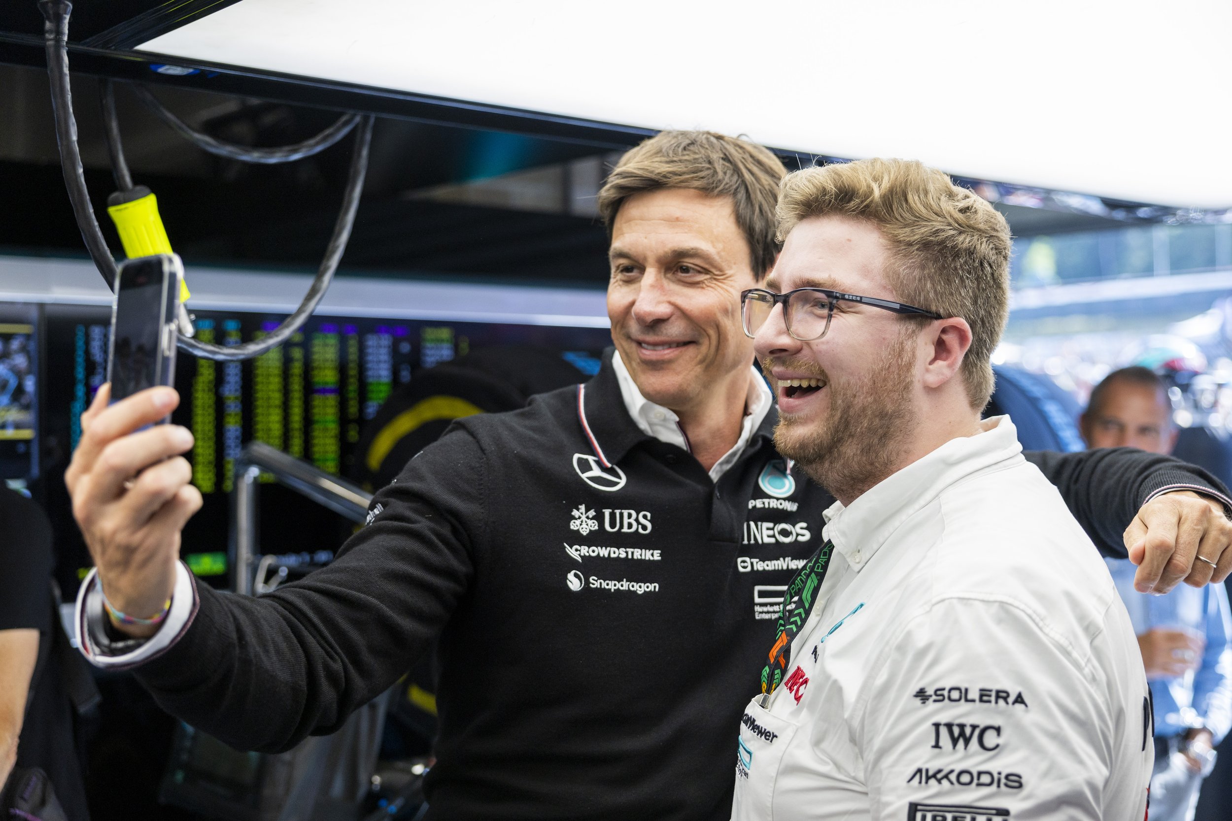 Toto Wolff and Jack Leslie, Toto in a black Mercedes jumper and  Jack in a white shirt, are taking a selfie together in a racing garage, with a backdrop of racing equipment and monitors.