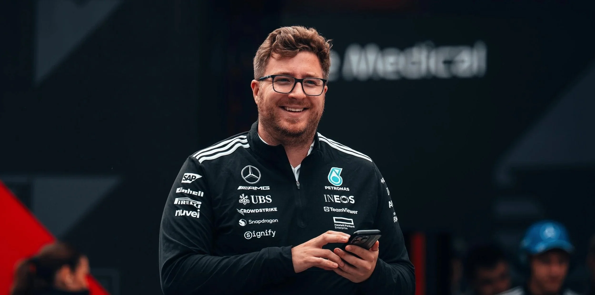 Jack Leslie with glasses smiling and holding a smartphone, wearing a black racing suit with sponsor logos, in a racing environment.