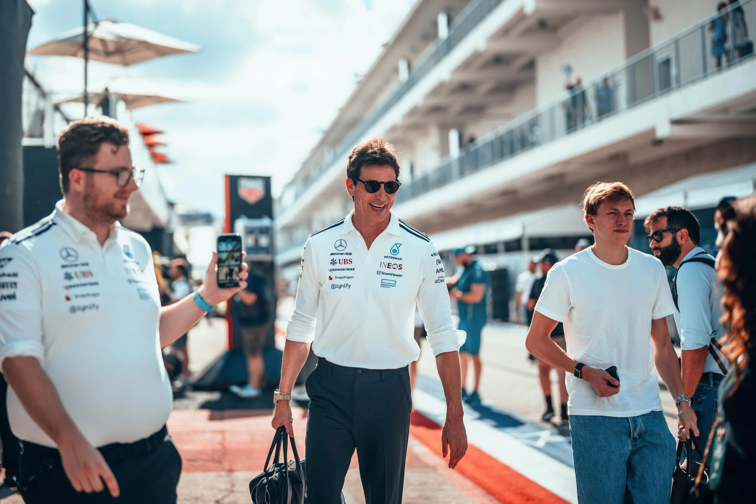 Toto Wolff and Jack Leslie walking through the F1 paddock. Jack films Toto and both are wearing white Mercedes tees.