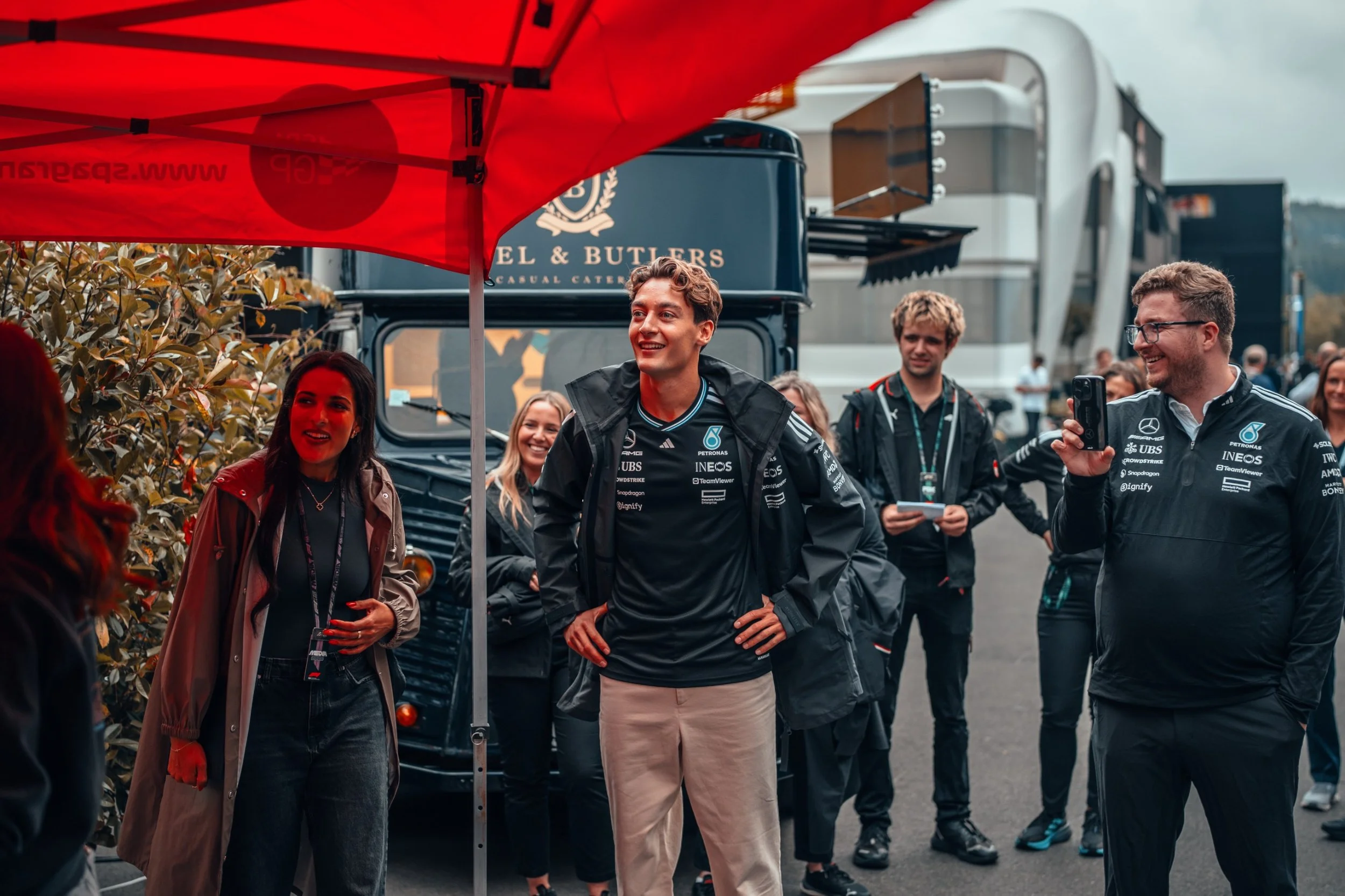 Group of people, some wearing team jackets, standing outdoors under a red canopy, engaging in conversation at a racing event with trucks and trailers in the background.