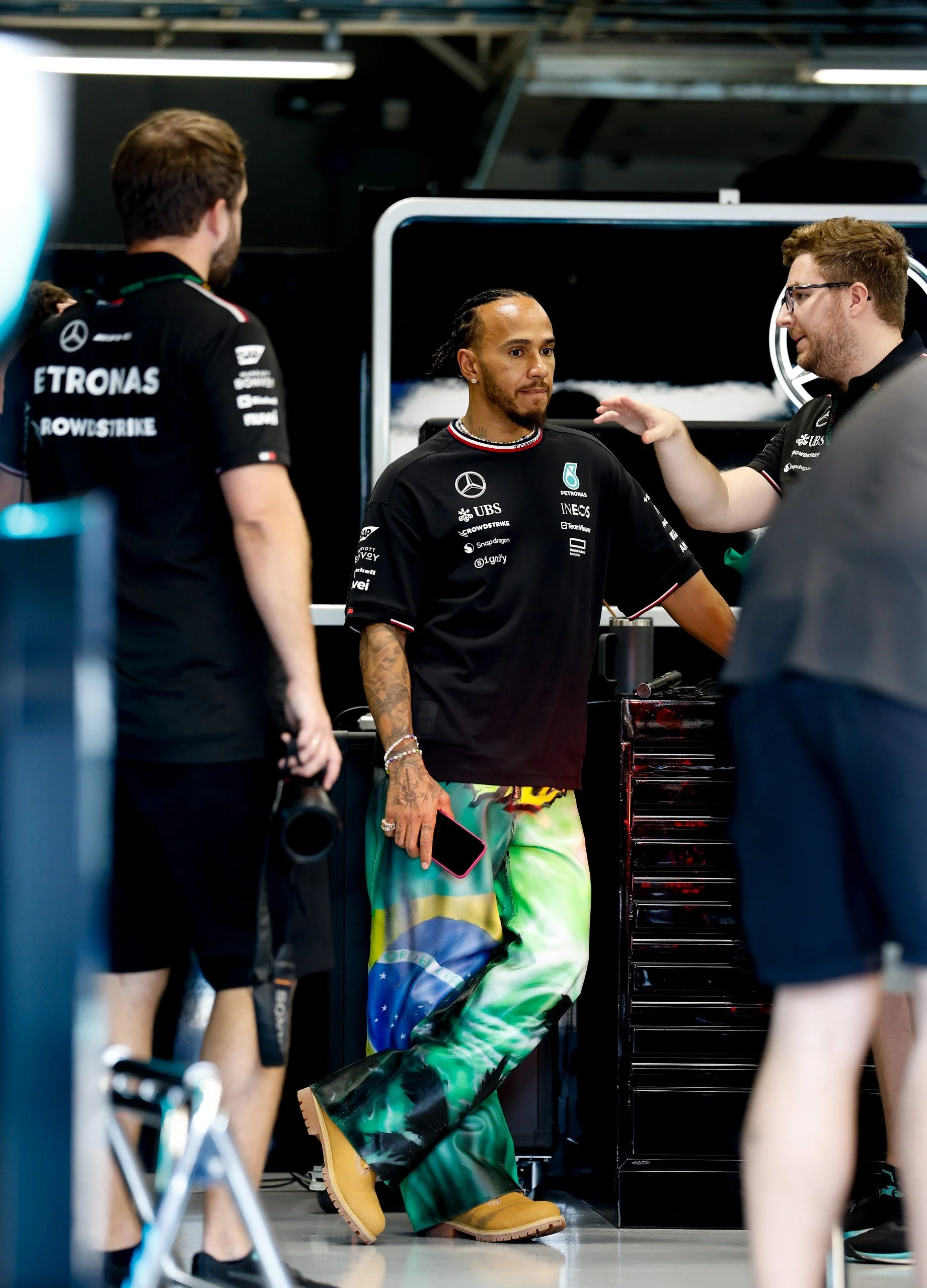 Lewis Hamilton, a Formula 1 driver, standing in a garage area with crew members around him. He is wearing colorful, patterned trousers and yellow boots, holding a phone in his right hand.