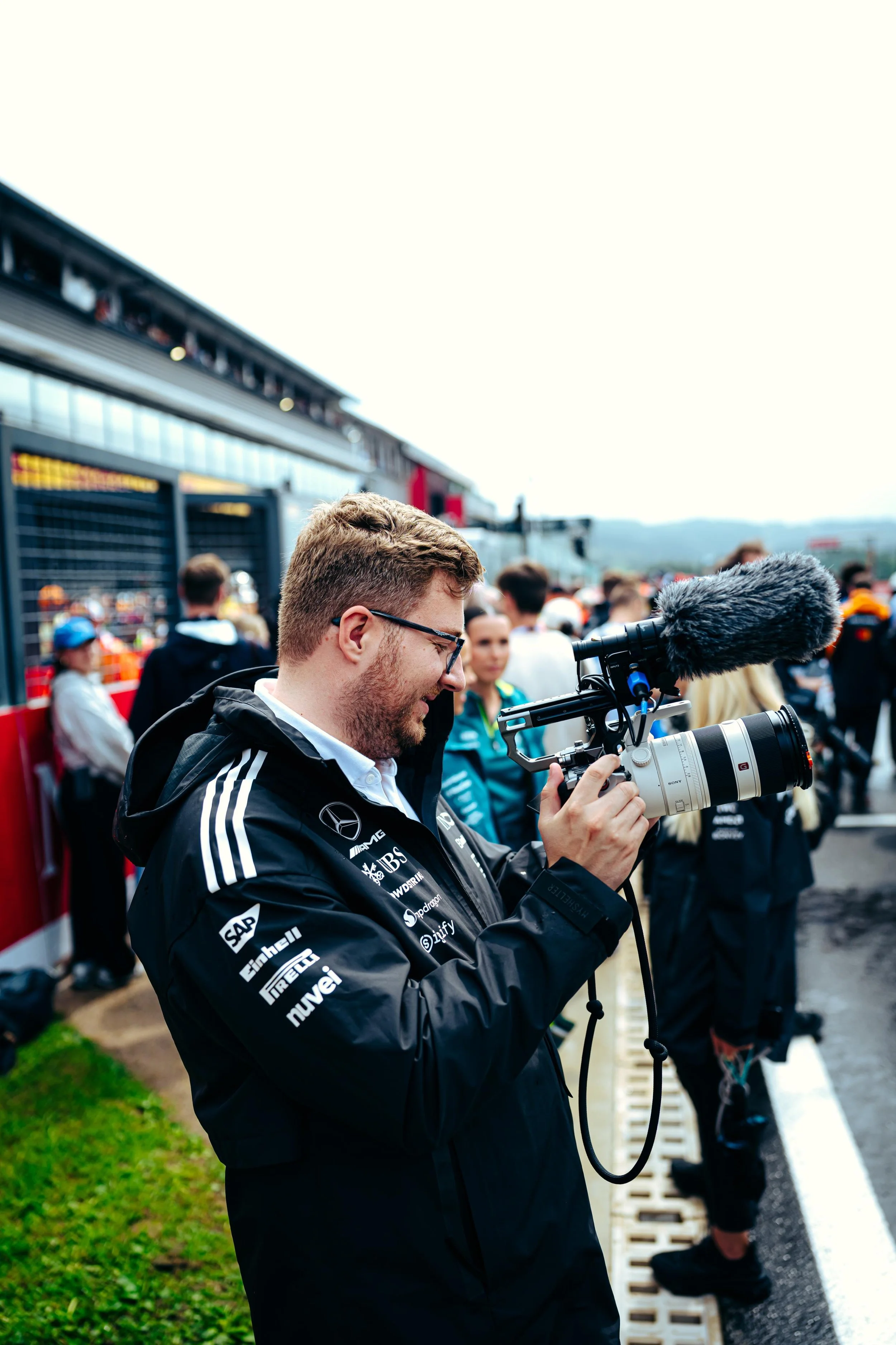 Jack Leslie with glasses holding a professional video camera with a microphone attached, standing among other people at what appears to be a race event or racing pit lane.