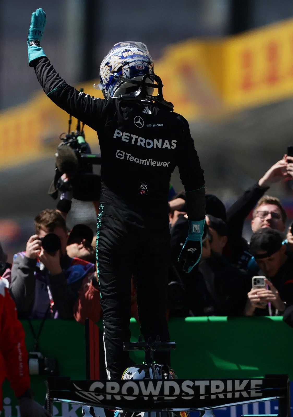 F1 driver George Russell in racing suit and helmet standing on his car, raising his fist in victory, surrounded by spectators at a race track.