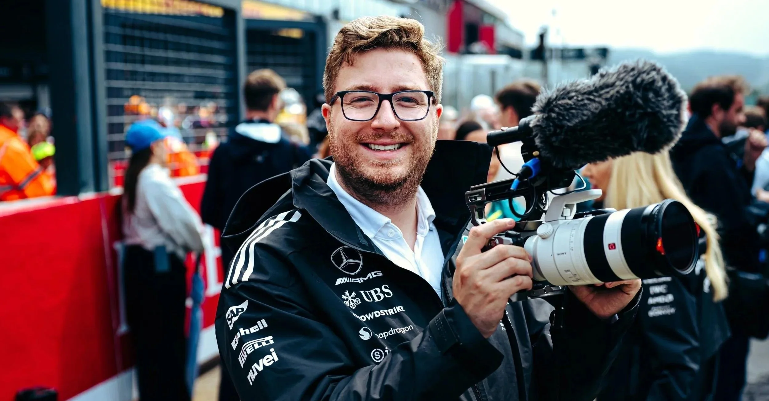 Jack Leslie with glasses and a beard, wearing a black racing jacket with sponsor logos, is smiling and holding a video camera. He is at a race event with a crowd of people in the background.