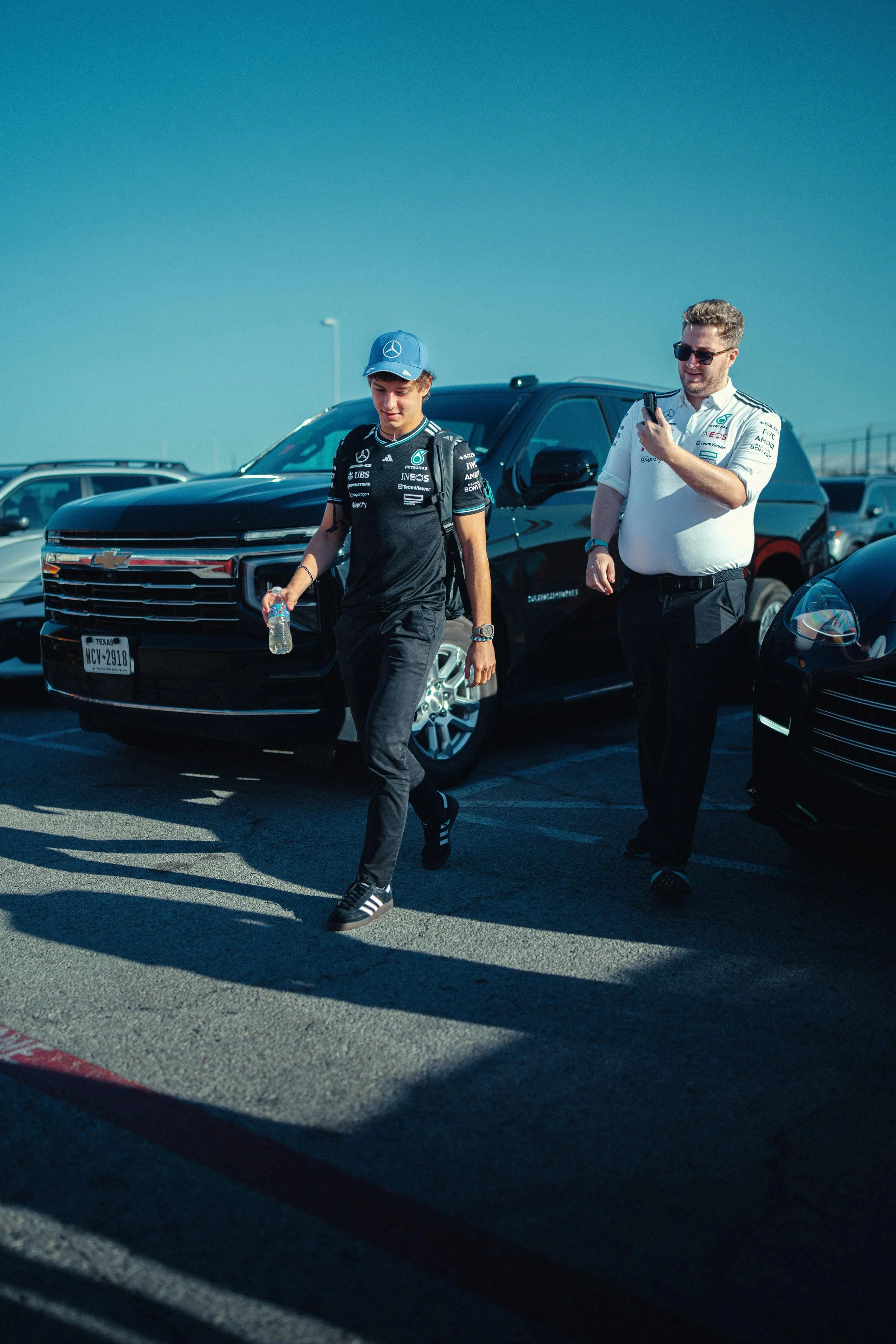 Kimi Antonelli and Jack Leslie walking in a parking lot, one wearing a black racing team shirt and cap with Mercedes logo, the other in a white racing shirt with sunglasses, looking at his phone, with parked cars behind them.
