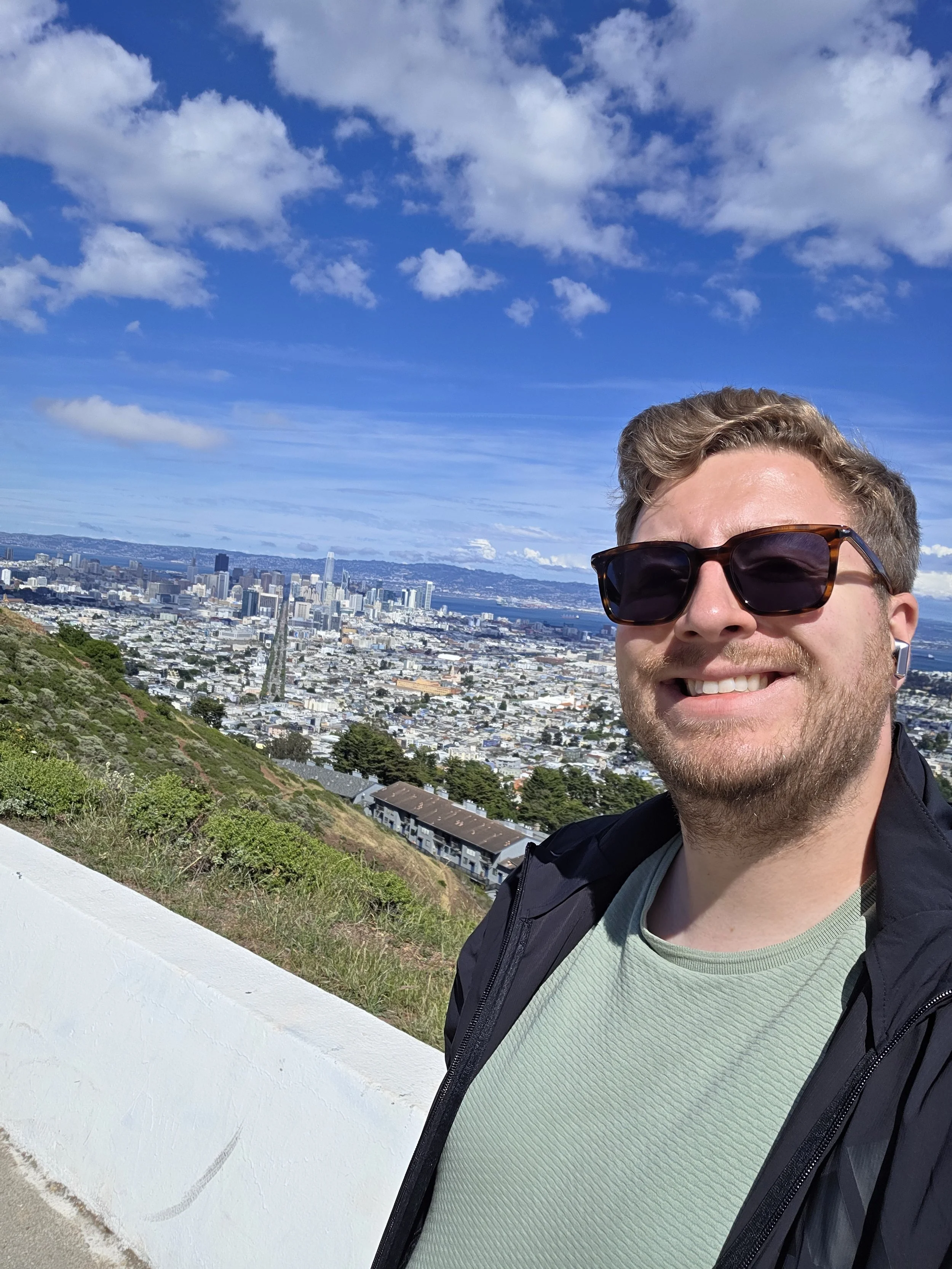 Jack Leslie smiling in sunglasses, standing on a hill overlooking a city skyline under a blue sky with scattered clouds.