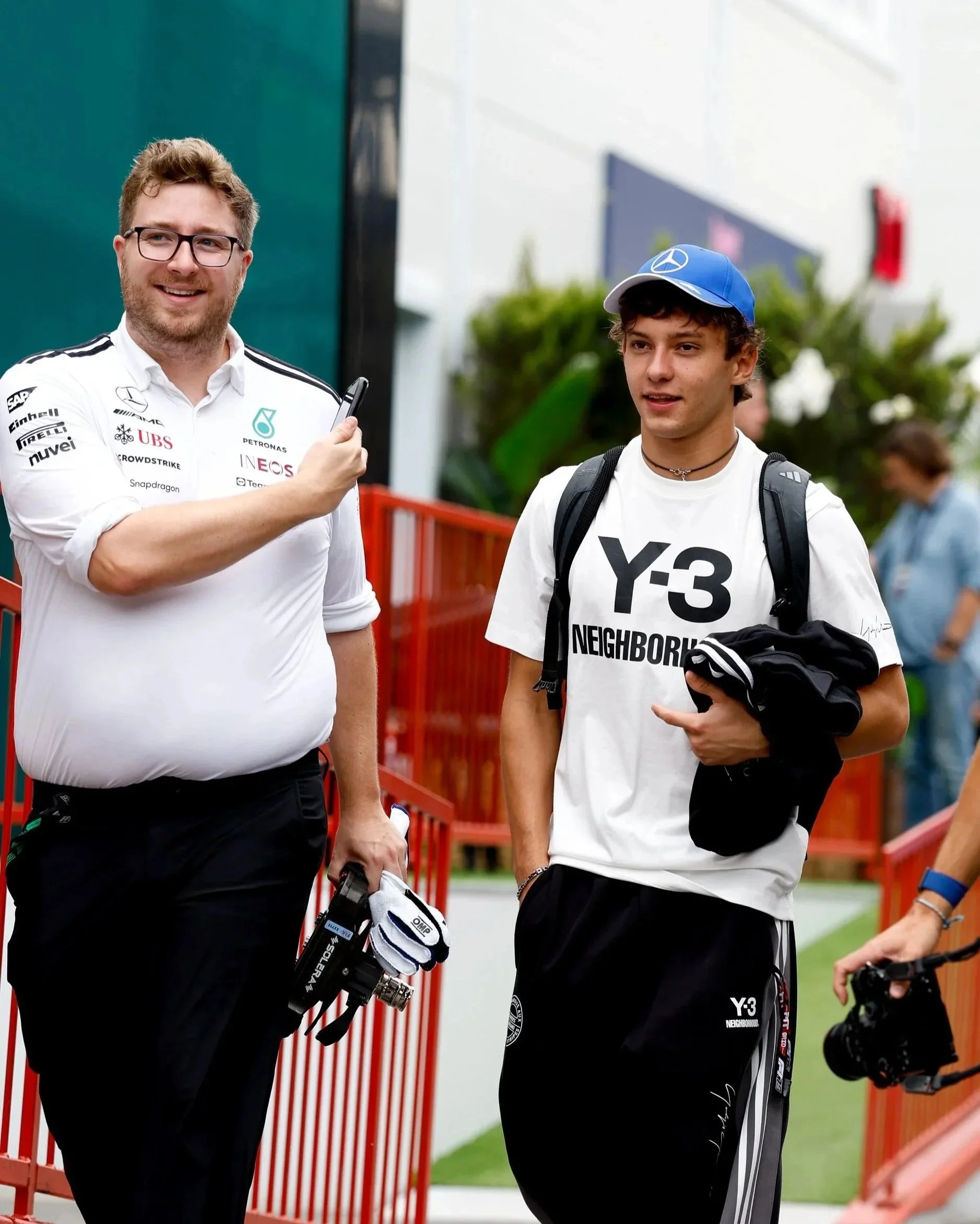 Jack Leslie and Kimi Antonelli walking. Jack wears a white shirt with sponsor logos, glasses, and holding a golf club. Kimi is wearing a white T-shirt with text, a blue cap with a logo, and carrying a backpack, holding a black jacket in his hand.