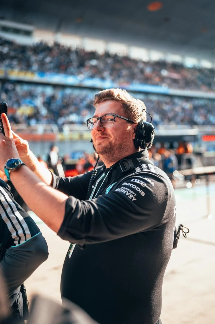 Jack Leslie with glasses and headphones taking a selfie in an F1 pit lane.