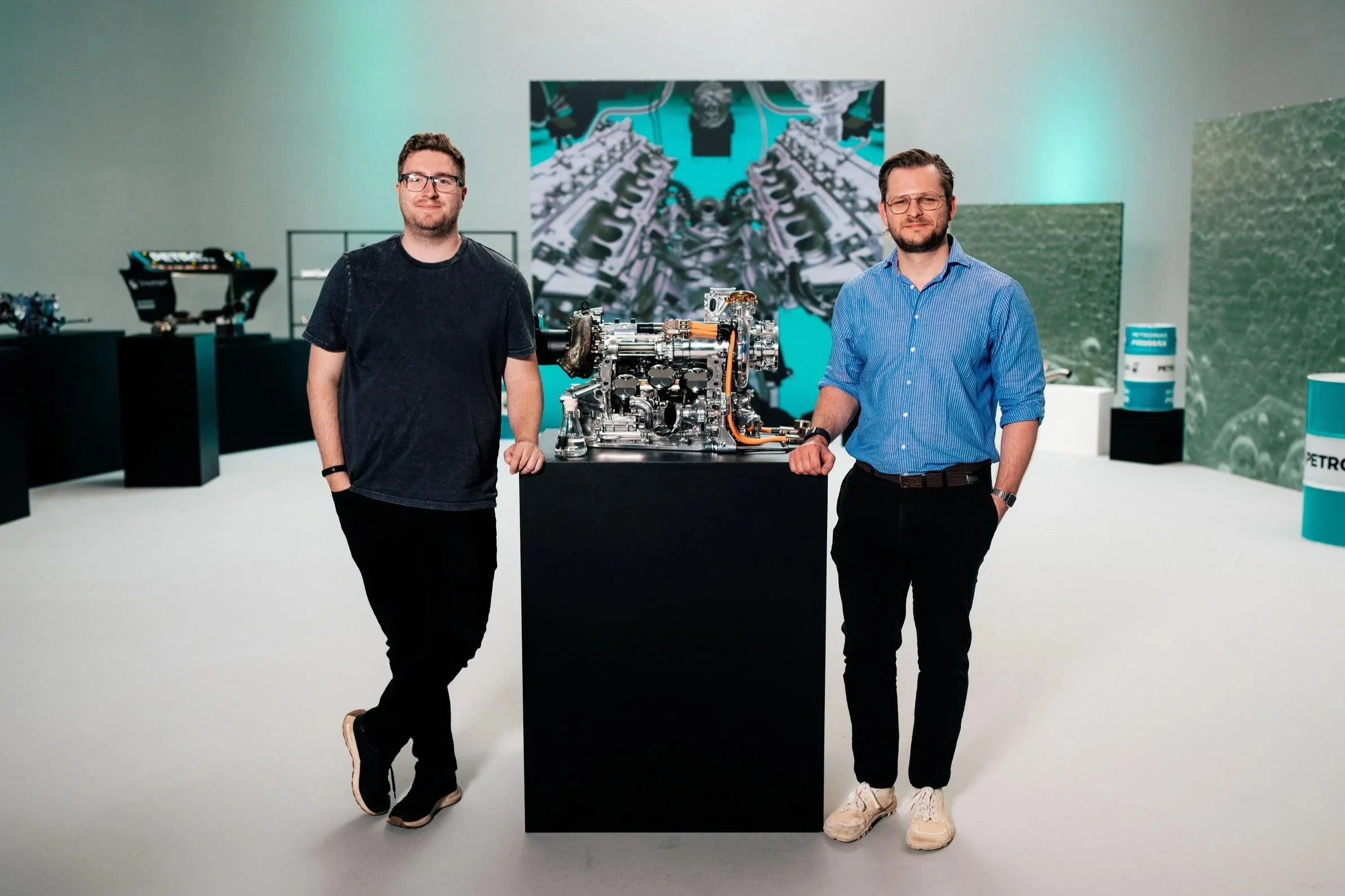 Two men standing on either side of a large mechanical object on display in a gallery. The man on the left is wearing a black t-shirt and glasses, and the man on the right is wearing a blue shirt and glasses. Behind them, there are various artwork and display cases, with a large digital screen displaying an animated image.