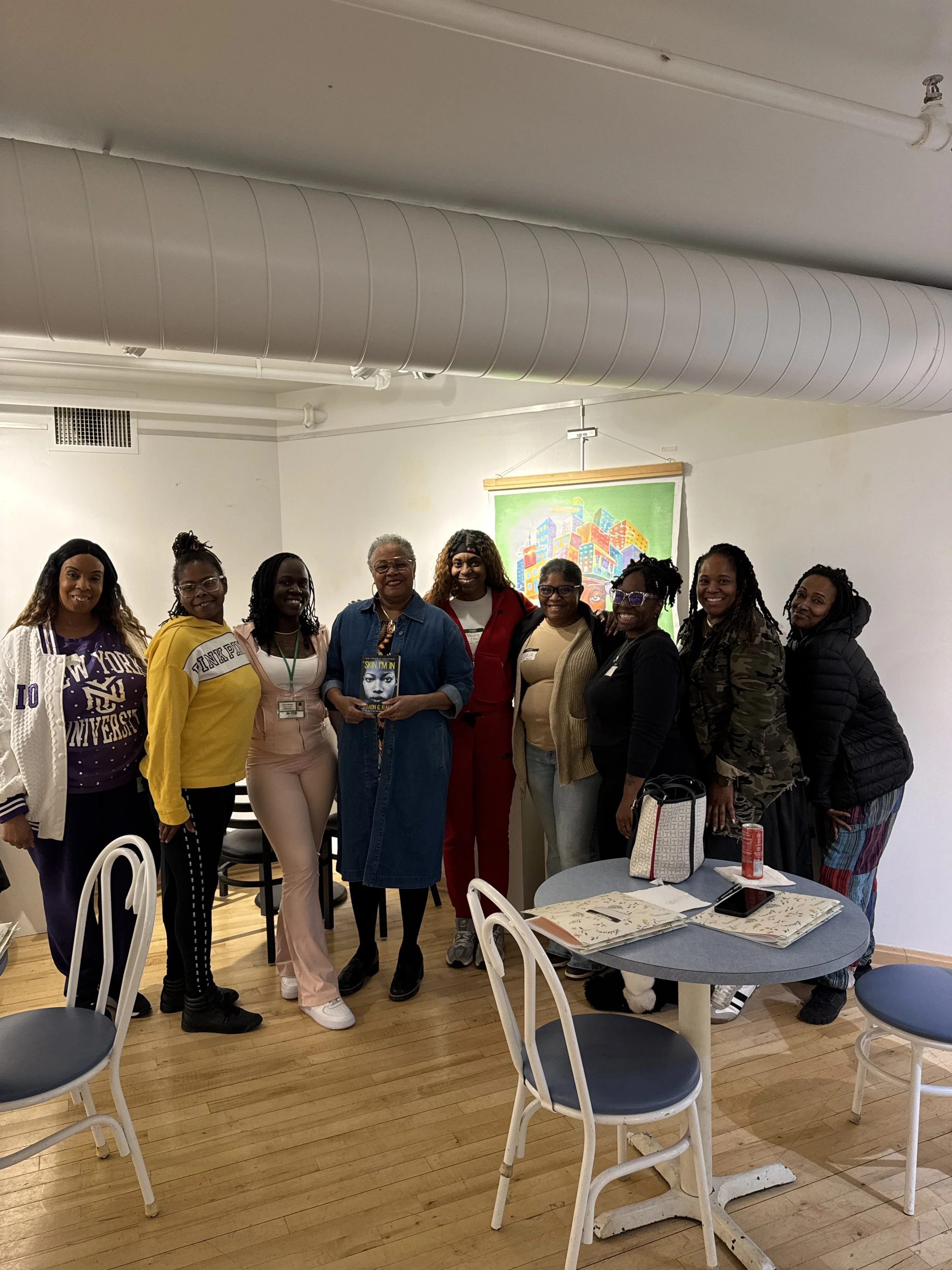A diverse group of ten women standing together indoors, smiling at the camera, with a stack of tables and chairs nearby and a colorful cityscape art piece in the background.