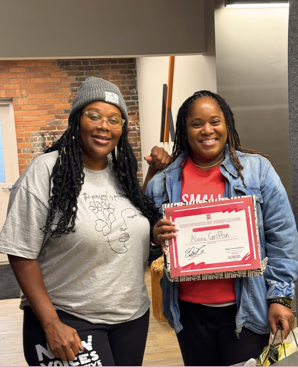 Two women standing indoors, one holding a certificate of completion and smiling, the other smiling and making a fist gesture.