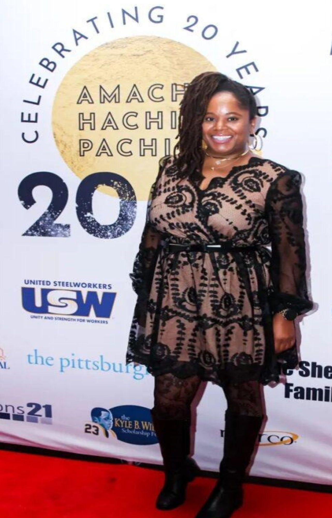 A woman standing on a red carpet at a celebration event, smiling at the camera. Behind her is a backdrop with logos and text, including 'Celebrating 20 Years,' 'AMACHI HACHI PACHI,' and 'USW.' She is wearing a patterned beige and black dress with lace details and black shoes.