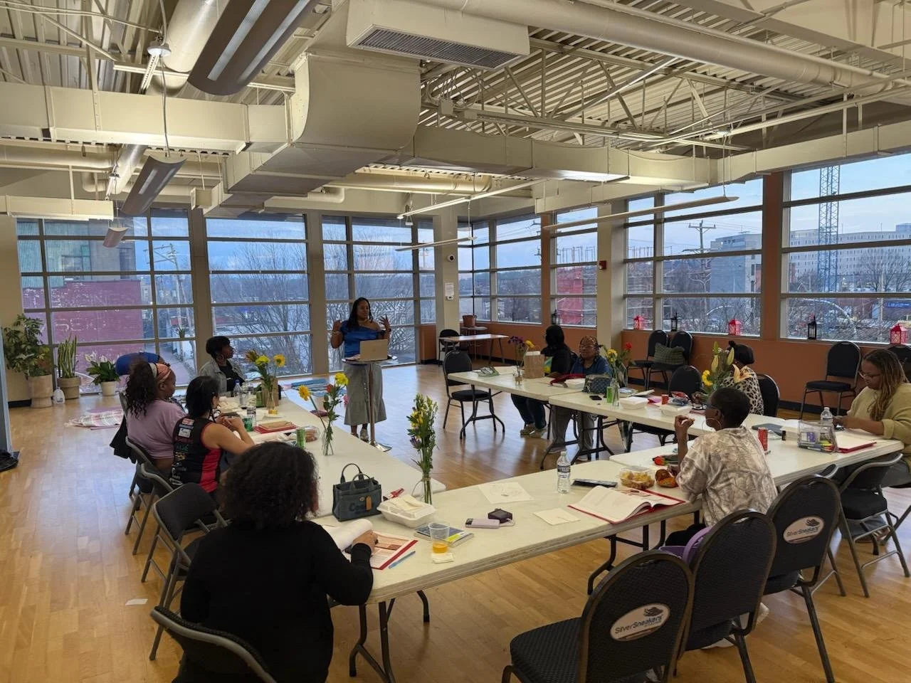 A woman stands in front of a group of people sitting around a large U-shaped table in a bright conference room with large windows. The attendees are listening and taking notes, with beverages and notebooks on the table. Flowers in vases are also on the table.
