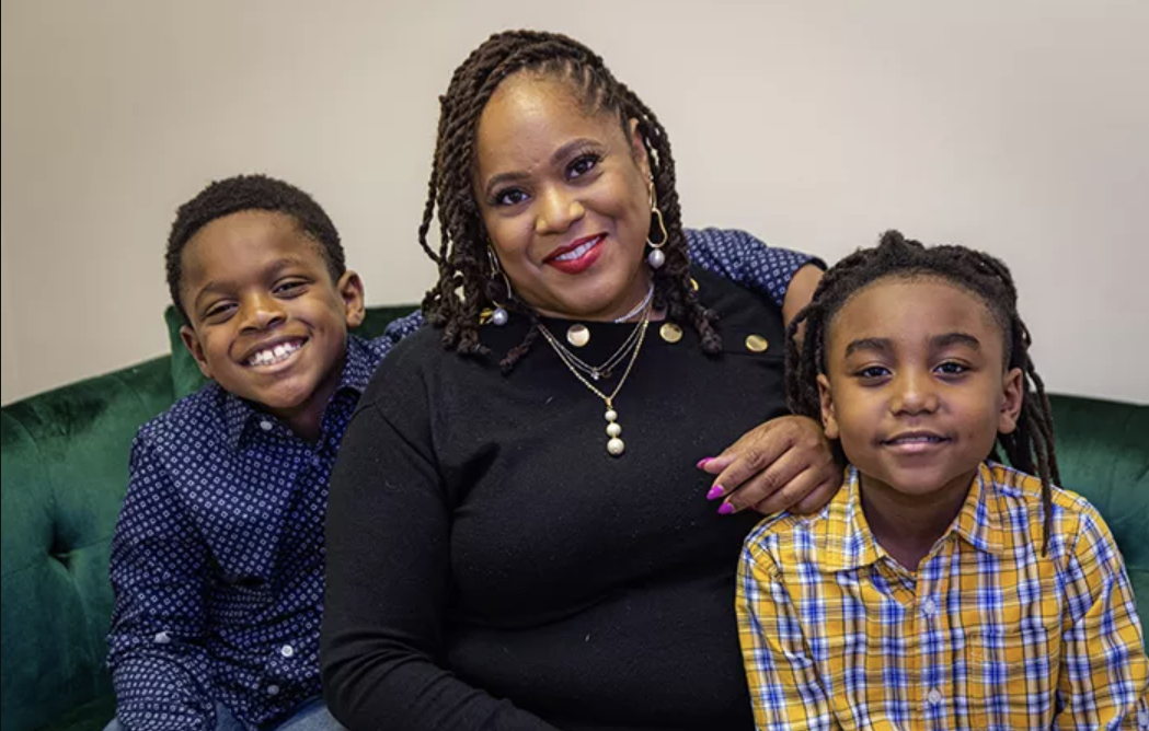 A woman with braids and red lipstick sitting on a green couch with two young boys, smiling at the camera. The boy on the left is wearing a blue patterned shirt, and the boy on the right is wearing a yellow plaid shirt.