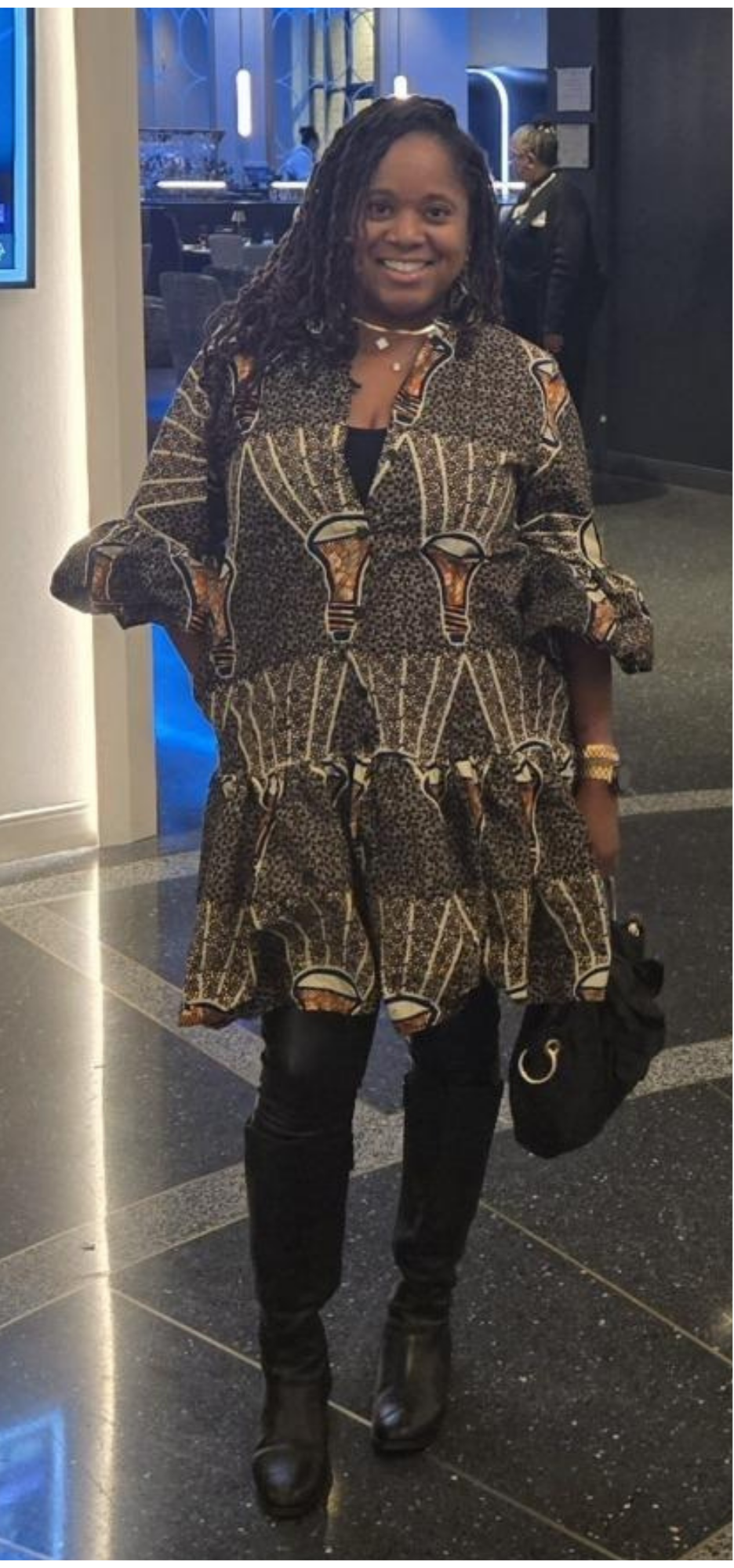 Woman smiling in a patterned dress, wearing black pants and boots, holding a black purse, standing in a modern indoor space.