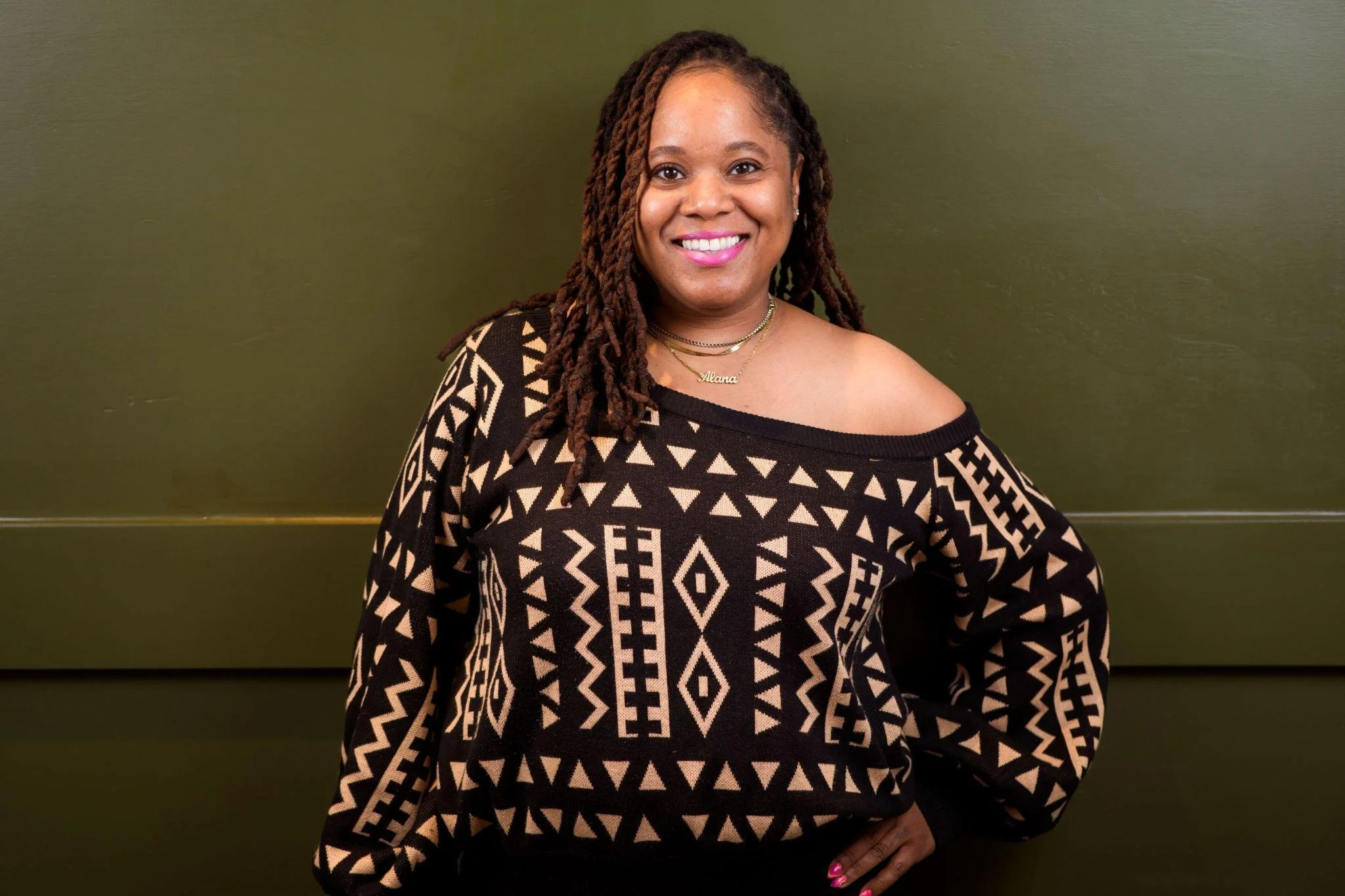A woman with long, twisted hair wearing a black and beige patterned off-shoulder top and gold jewelry, smiling in front of an olive green wall.