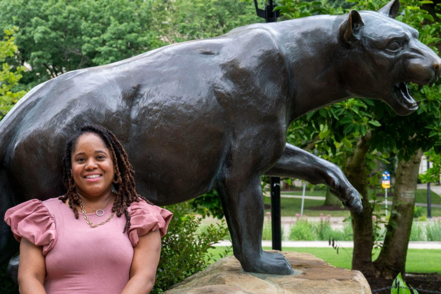 A woman with curly hair and jewelry sitting on a rock in front of a large bronze panther statue in a green outdoor park setting.