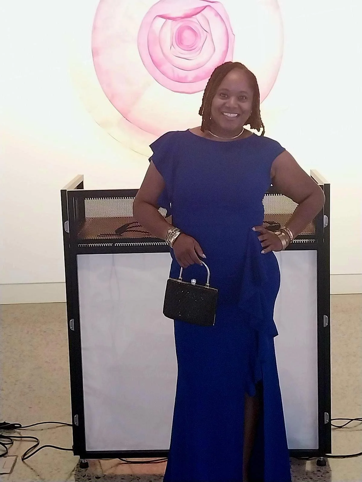 A woman in a royal blue dress standing in front of an abstract pink and purple floral artwork, holding a black clutch purse, smiling.