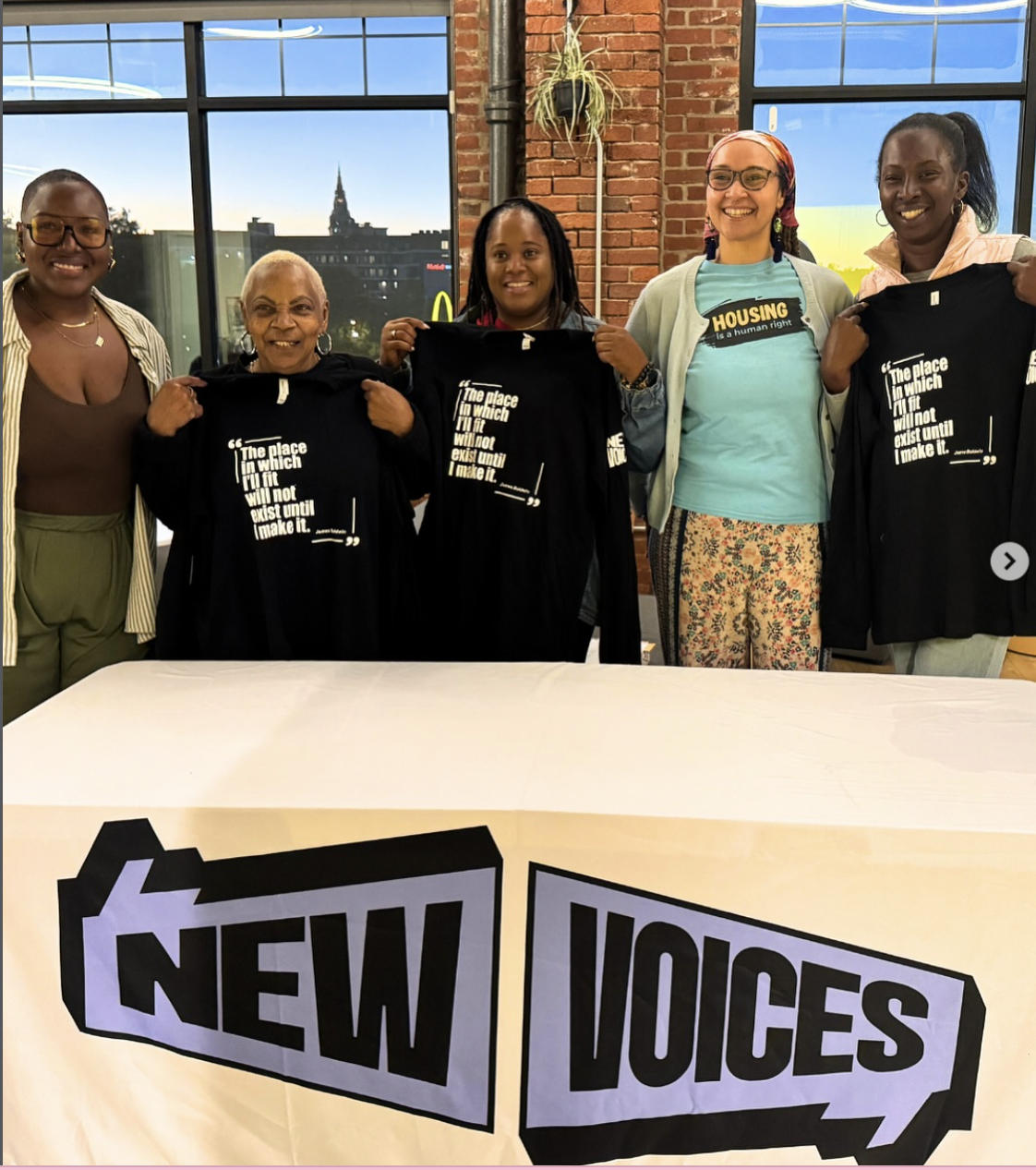 Five women standing behind a table with a sign that reads 'New Voices' in bold black letters. The women are holding black t-shirts with writing on them, smiling in a room with large windows showing a cityscape and a sunset.