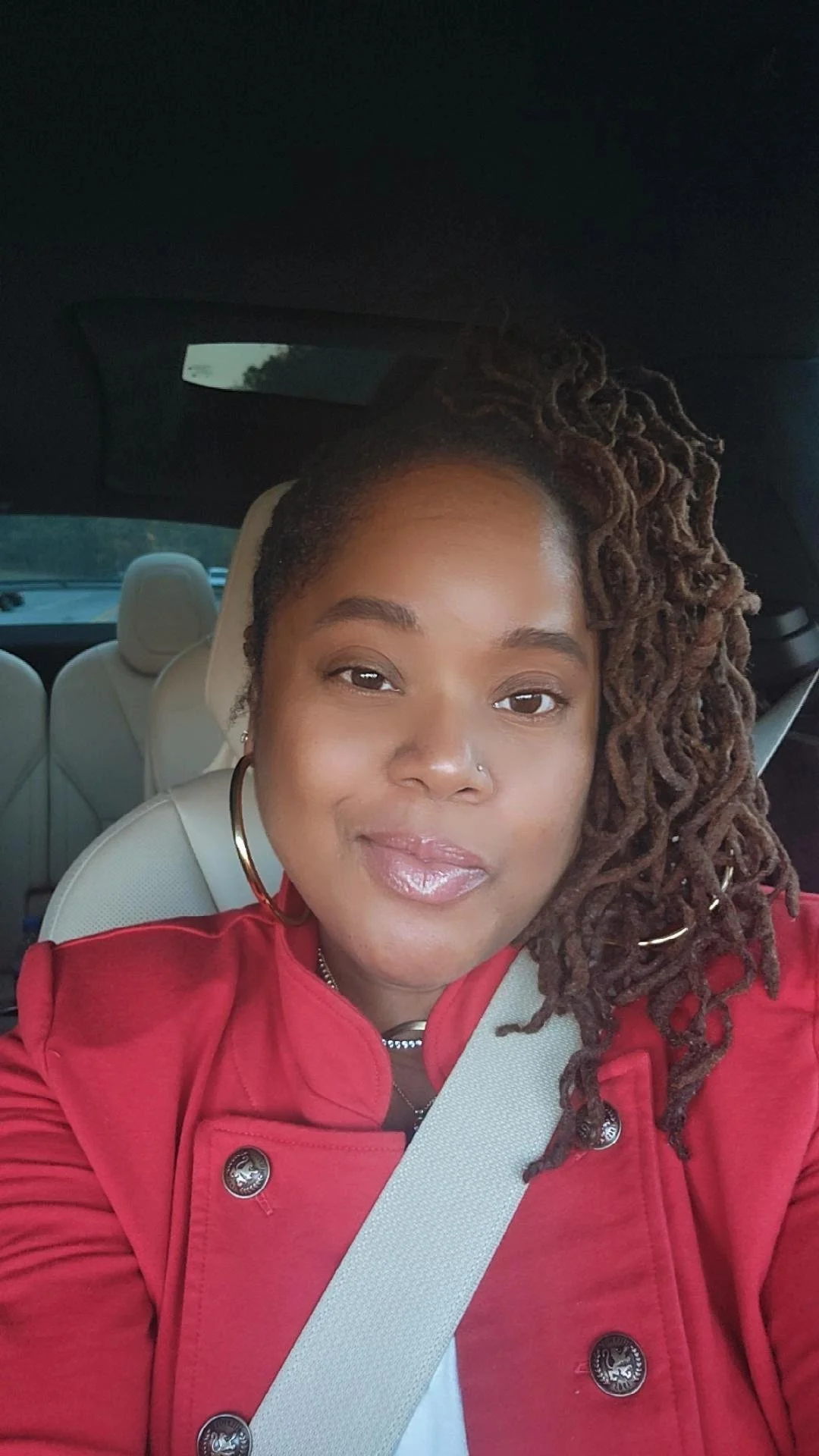 A woman with styled dreadlocks, wearing a red jacket and large hoop earrings, sitting in a car with beige interior, taking a selfie.