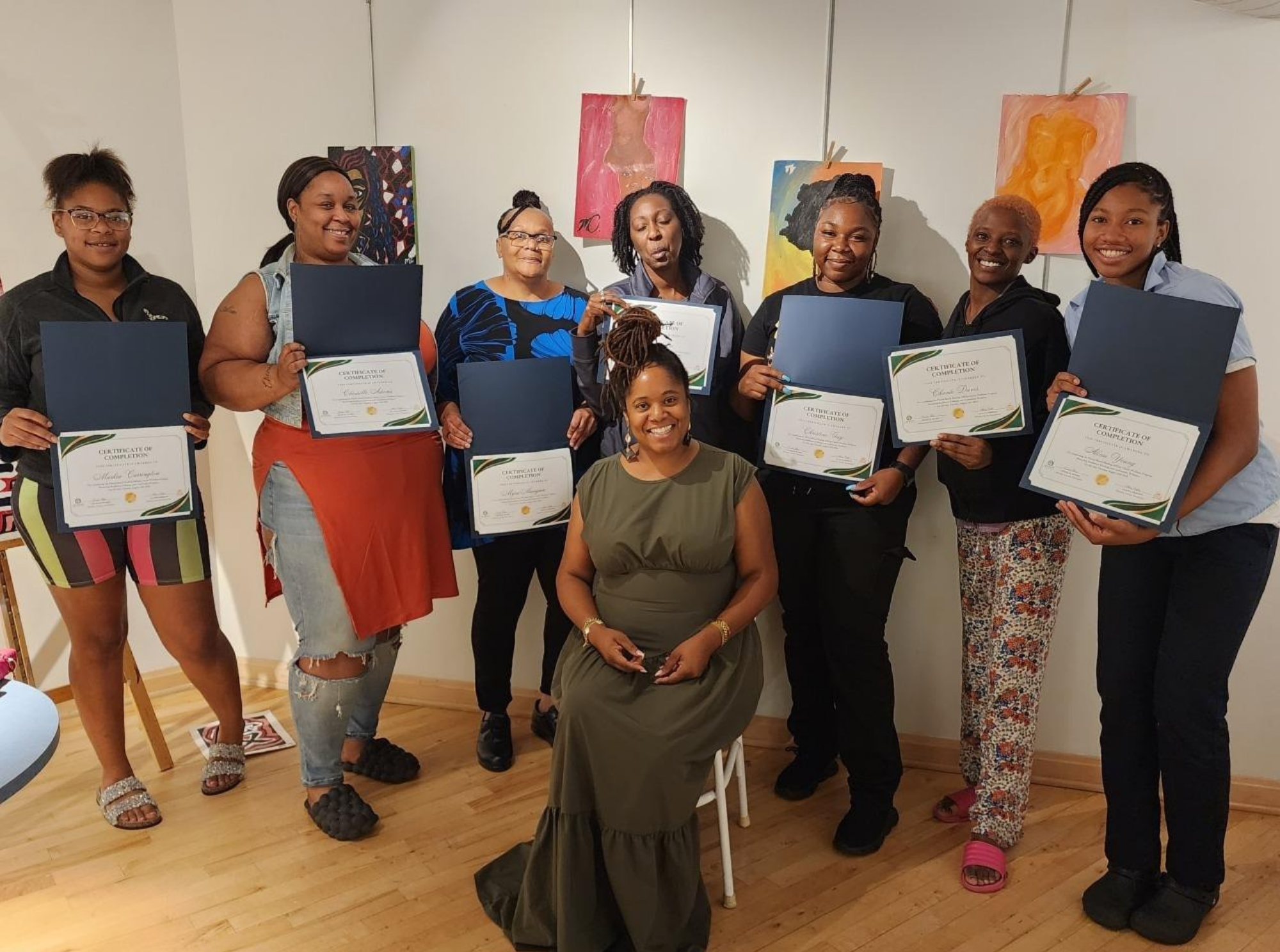 A group of women, some holding certificates, gathered in front of a white wall with colorful abstract paintings, celebrating an achievement or graduation in a warmly lit indoor space with wooden flooring.