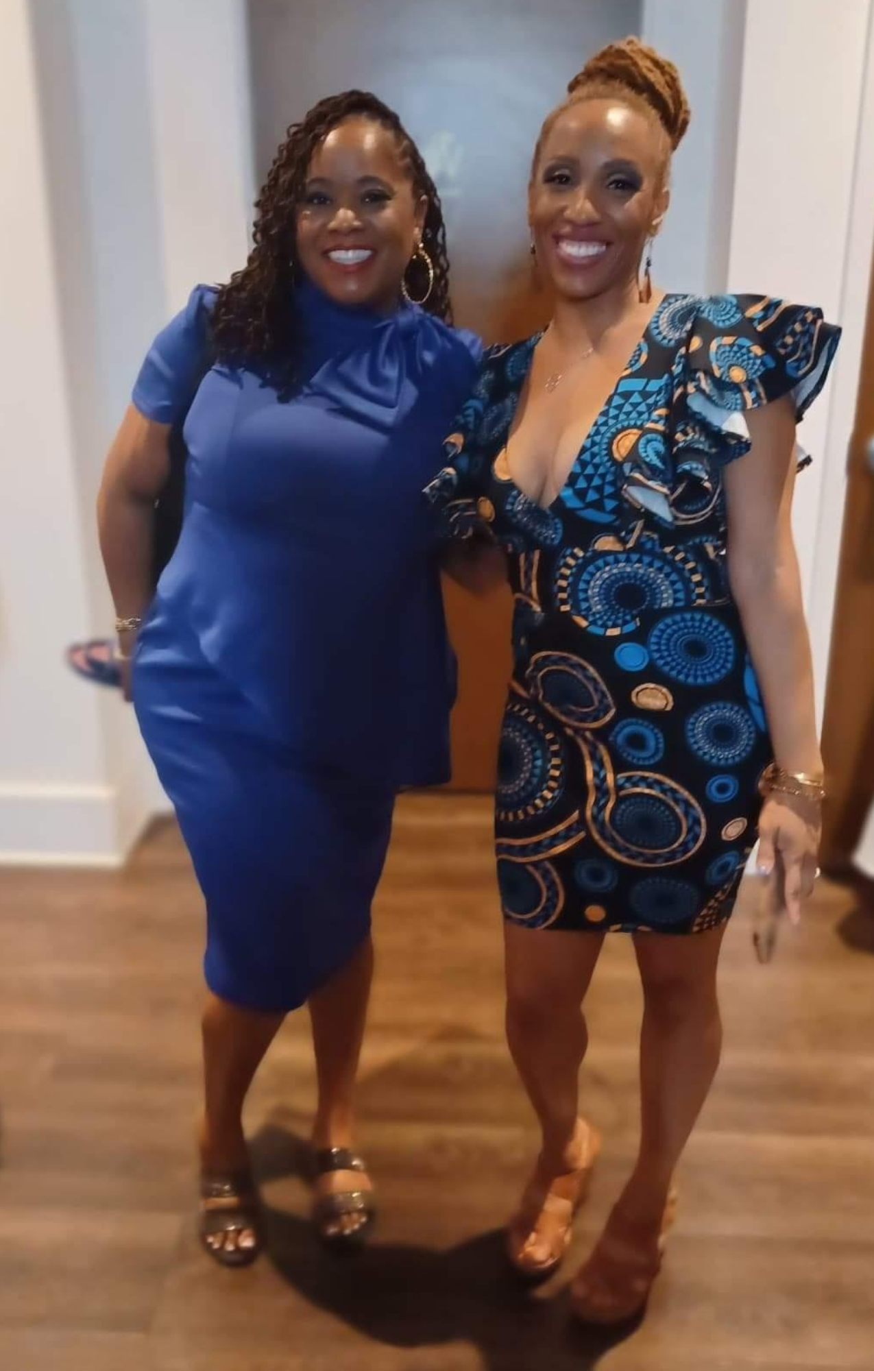 Two women standing indoors, smiling, dressed in elegant blue and patterned dresses, wearing high heels and jewelry.