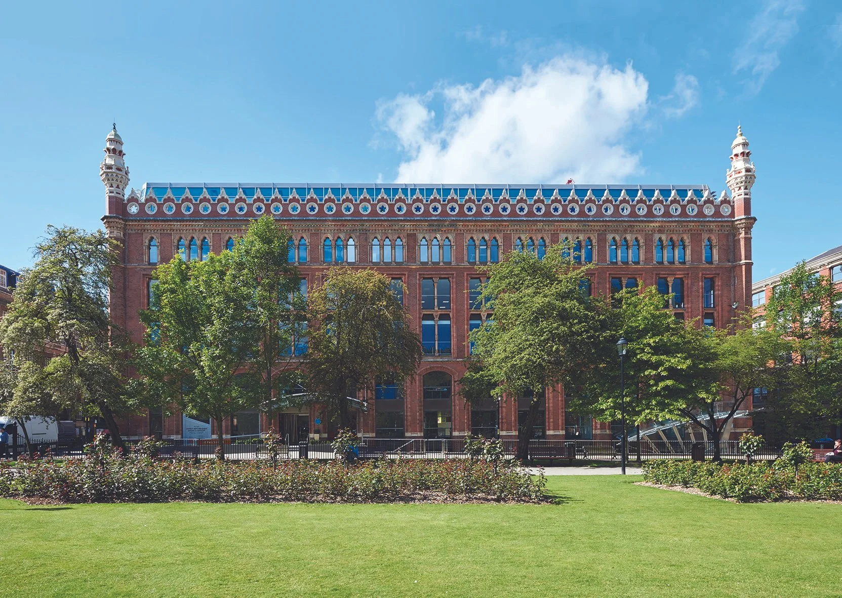 A historic brick building with decorative white accents and towers, surrounded by trees and a park with green grass under a blue sky with clouds.
