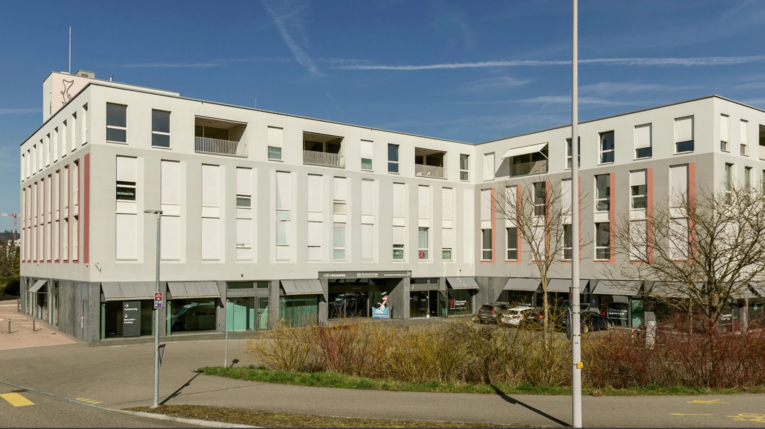 Moderne weiße sechsstöckige Gebäude mit großen Fenstern, Balkonen und Geschäftsläden im Erdgeschoss, unter blauem Himmel mit Wolkenformationen, Bäumen und Parkplatz im Vordergrund.