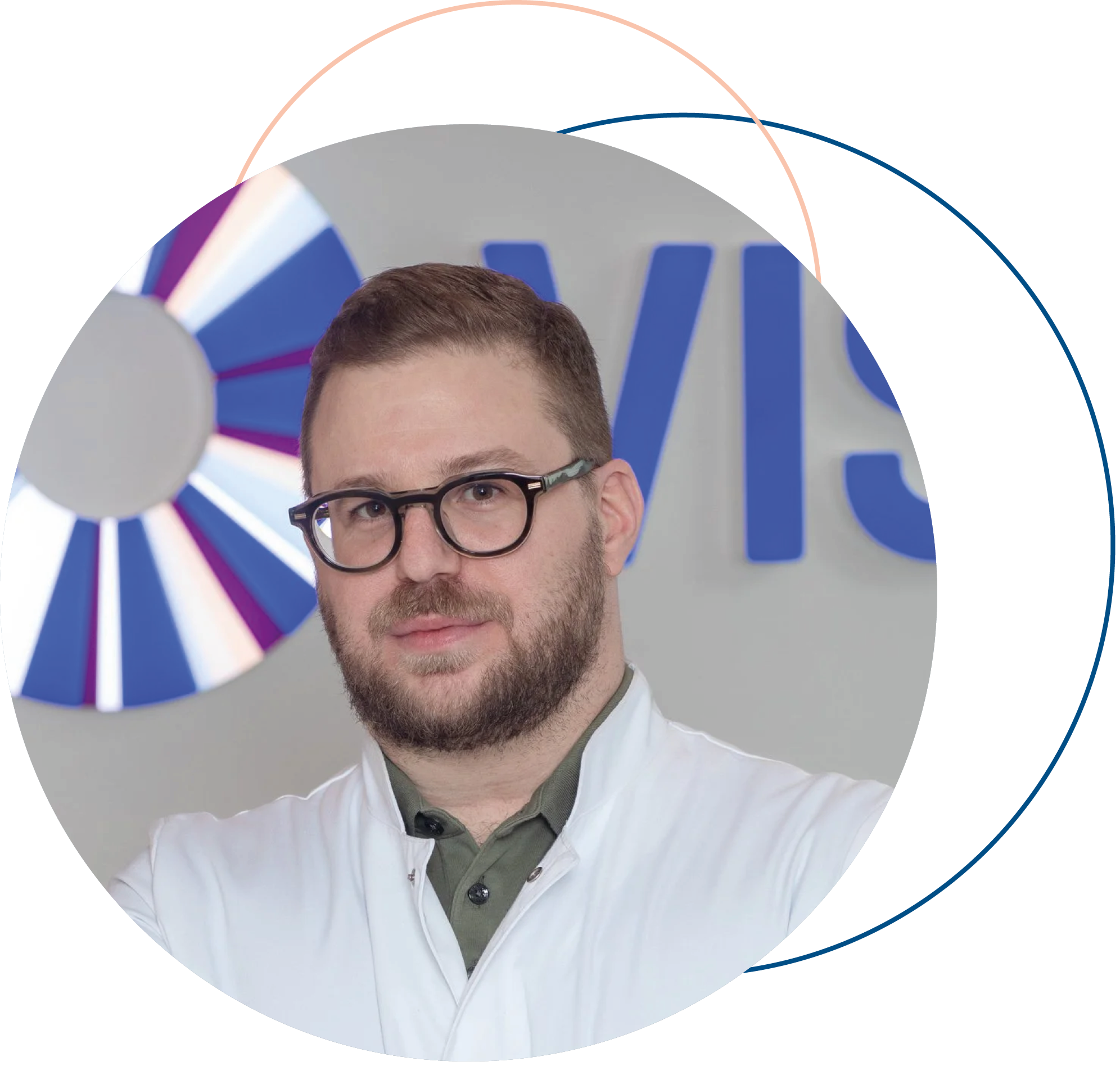Ein junger Mann mit kurz braunem Haar, Bart und Brille trägt eine weiße Jacke und steht vor einem Hintergrund mit einem blauen Logo und einem bunten Rad, vermutlich bei einer Veranstaltung oder in einem Studio.