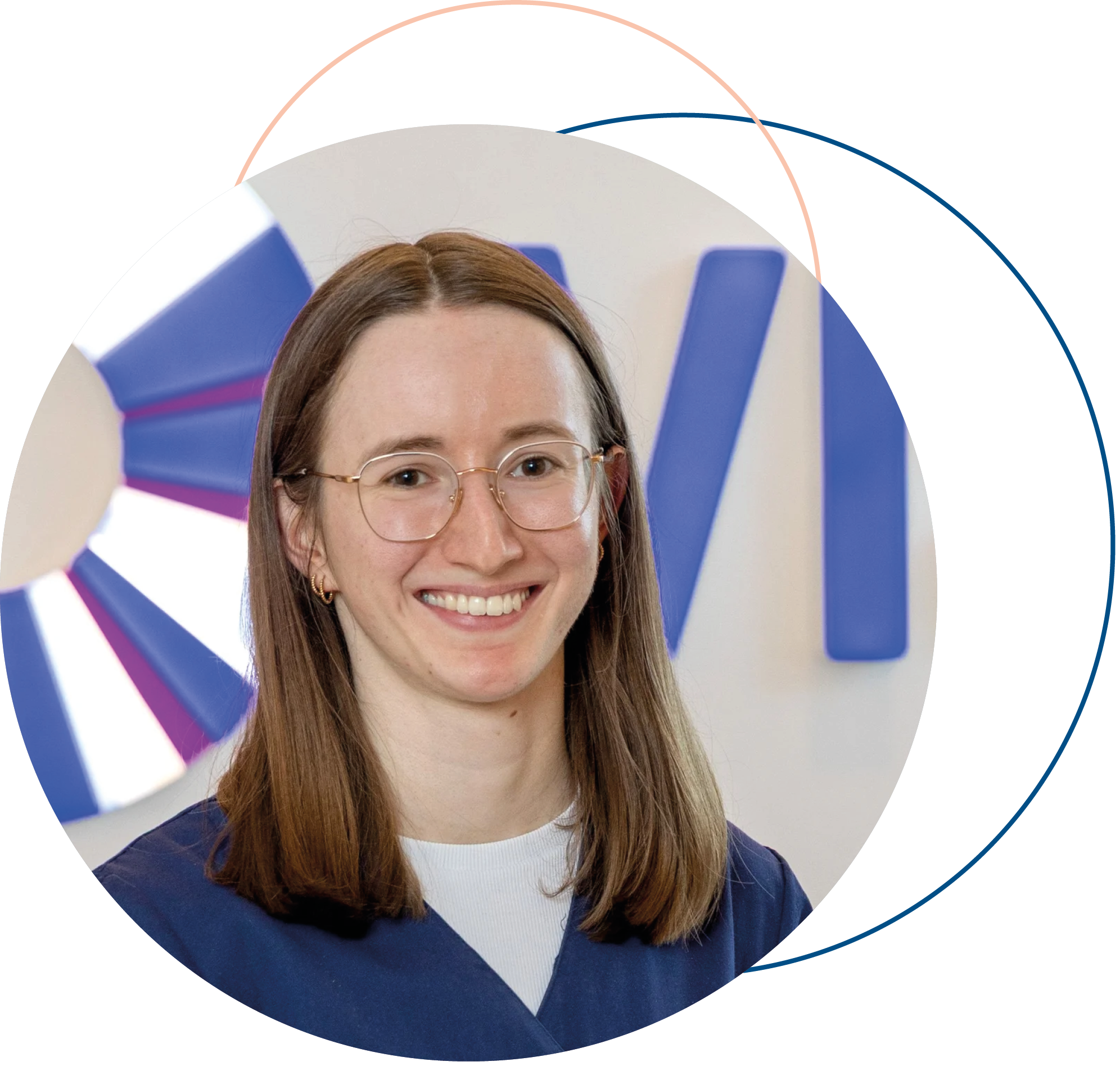 Junge Frau mit Brille, braune Haare, trägt einen blauen Blazer und ein weißes Hemd, lächelnd vor einer weißen Wand mit blaue und pinke Akzente.