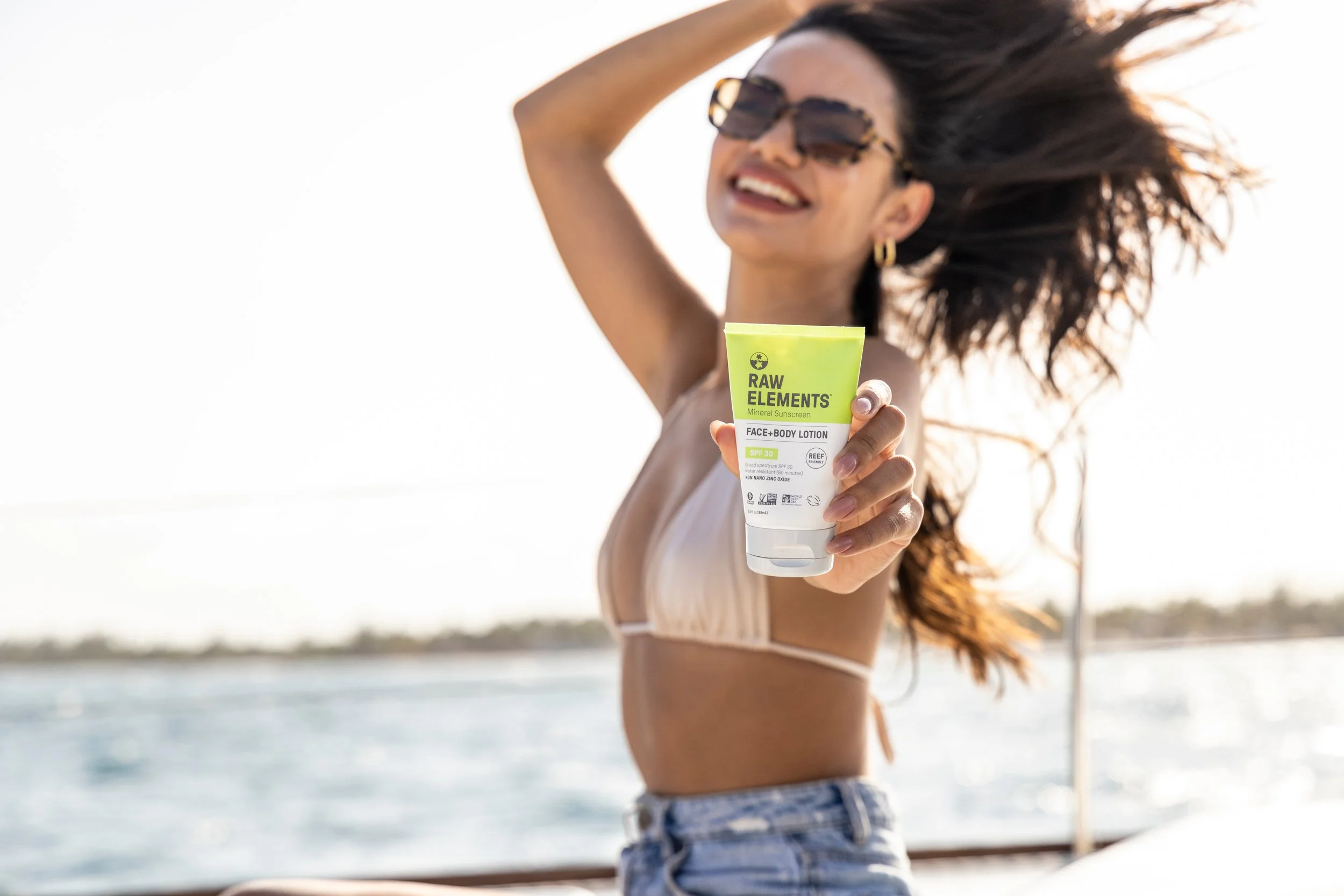 A woman with brown hair wearing sunglasses, a white swimsuit, and denim shorts at the beach, holding a tube of Raw Elements mineral sunscreen with SPF 30, smiling with her hair blowing in the wind.