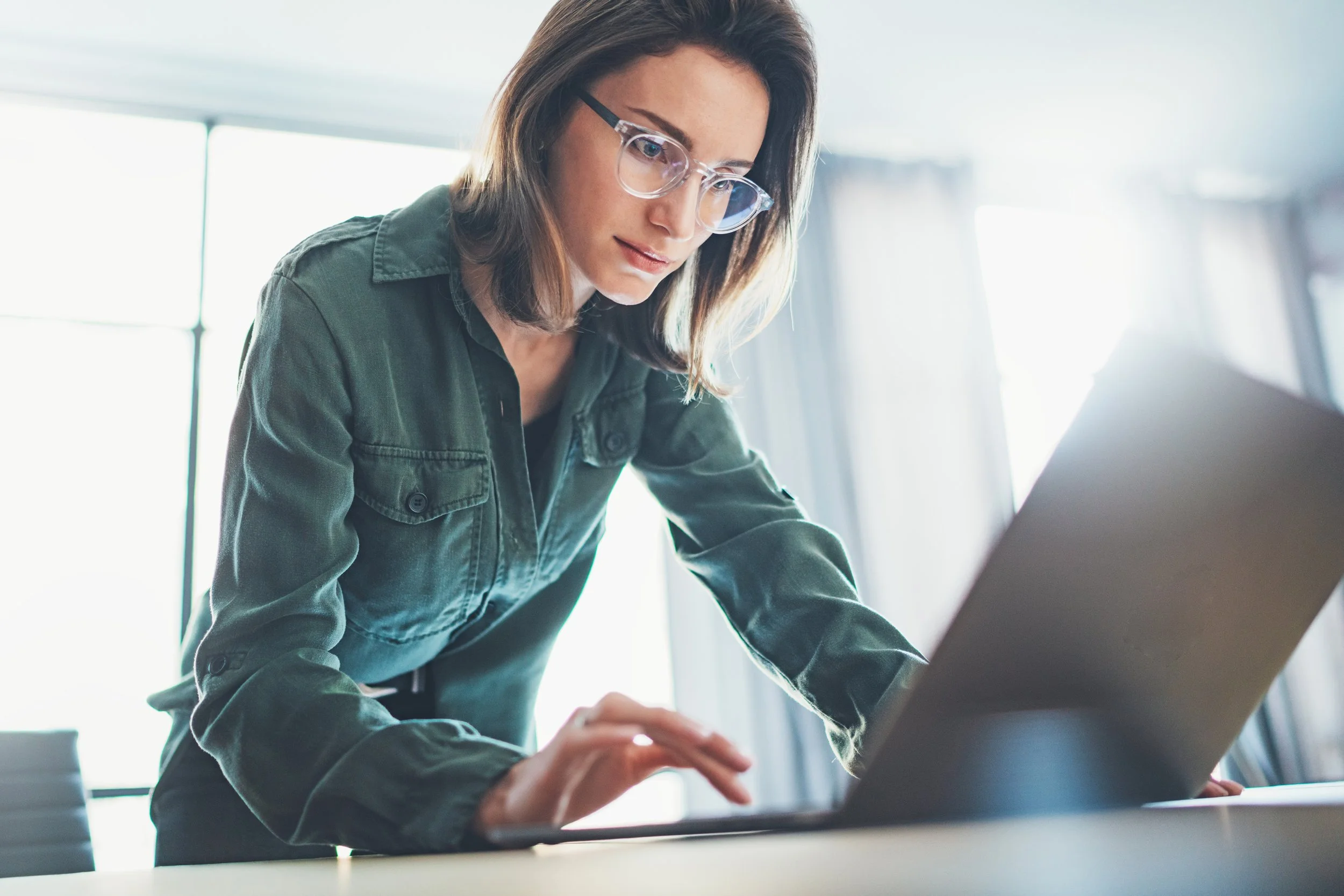 A woman with glasses and a green jacket working on a laptop in a bright room.