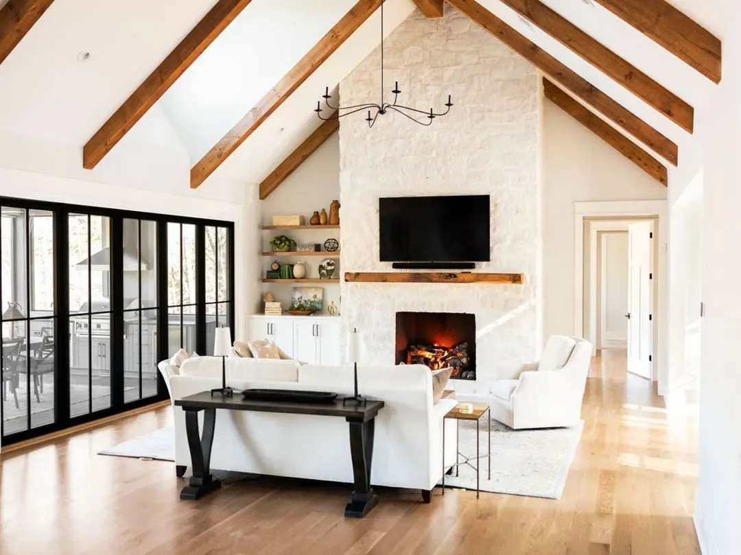 Living room with white walls, a stone fireplace with a mounted flat-screen TV above, wooden beams on the ceiling, and a black chandelier. There are white sofas, glass sliding doors leading to an outdoor deck, wood flooring, and built-in shelves with decorative items.