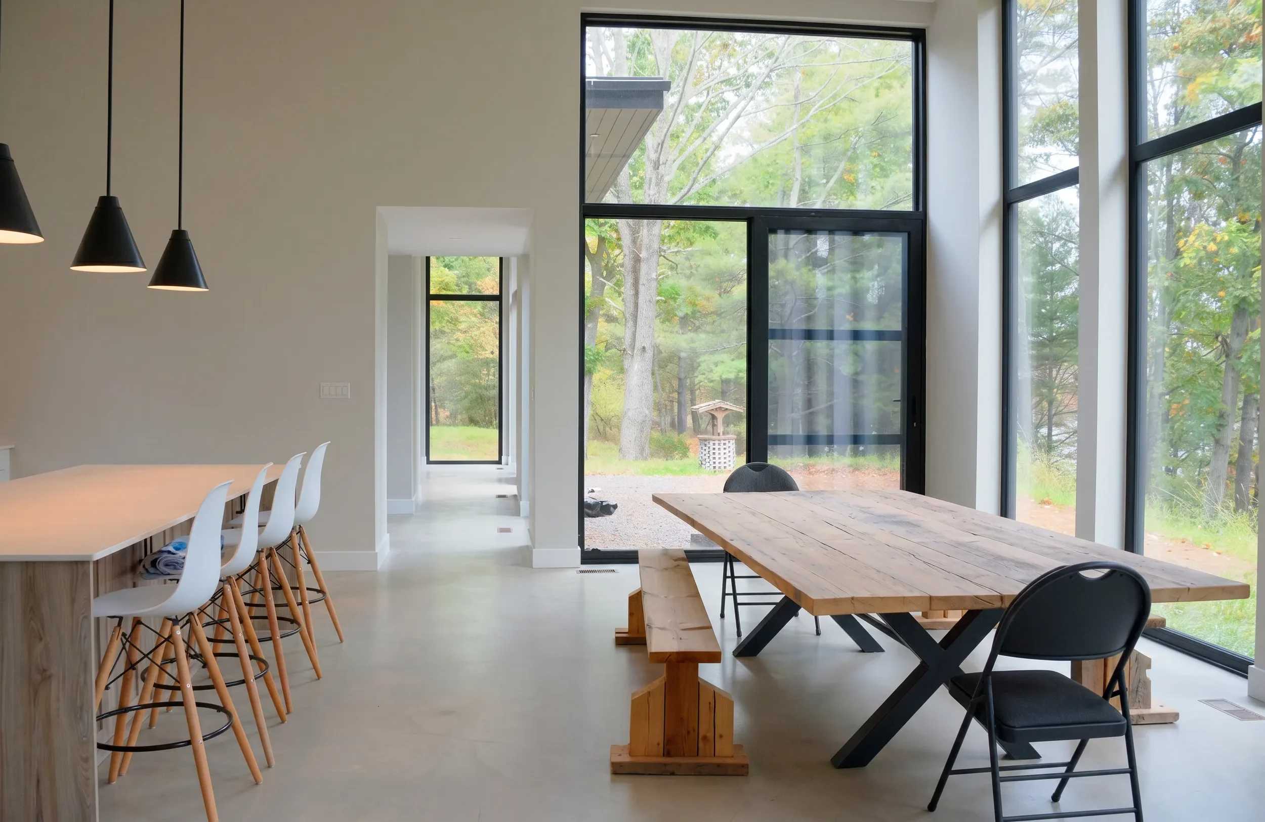 Interior of a modern home with large floor-to-ceiling windows showing a view of green trees outside. The room features a white dining table with white chairs, a wooden table with black legs, and black pendant lights hanging from the ceiling.