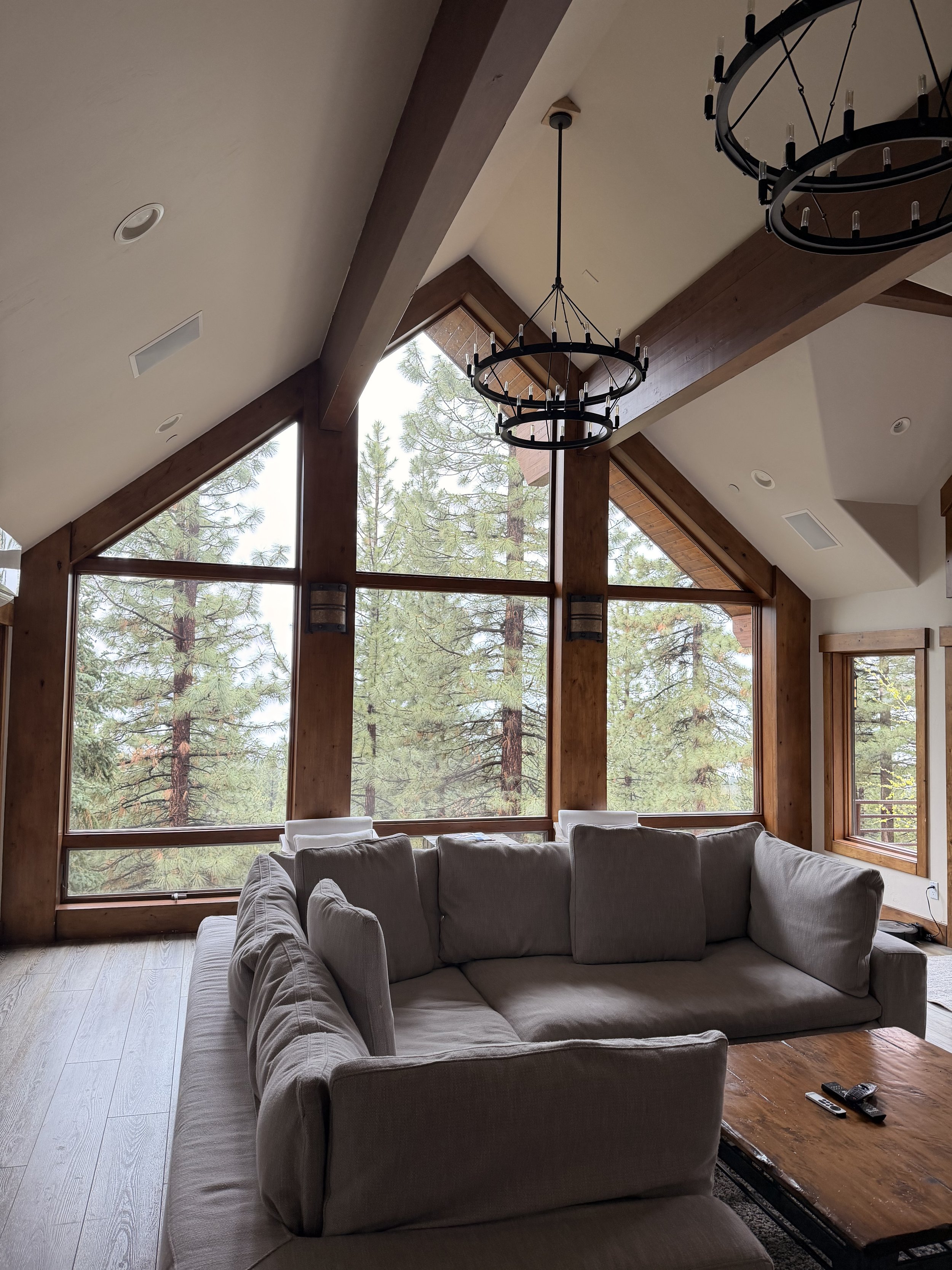 Living room with large windows showing a forest outside, two black circular chandeliers hanging from a vaulted ceiling, wooden beams, and a beige sectional sofa with a wooden coffee table and remote controls on it.