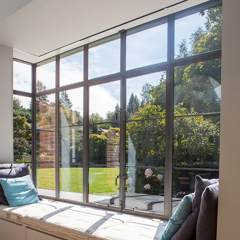 Sunlit indoor seating area with large glass windows offering a view of a green backyard with trees and a cloudy sky.