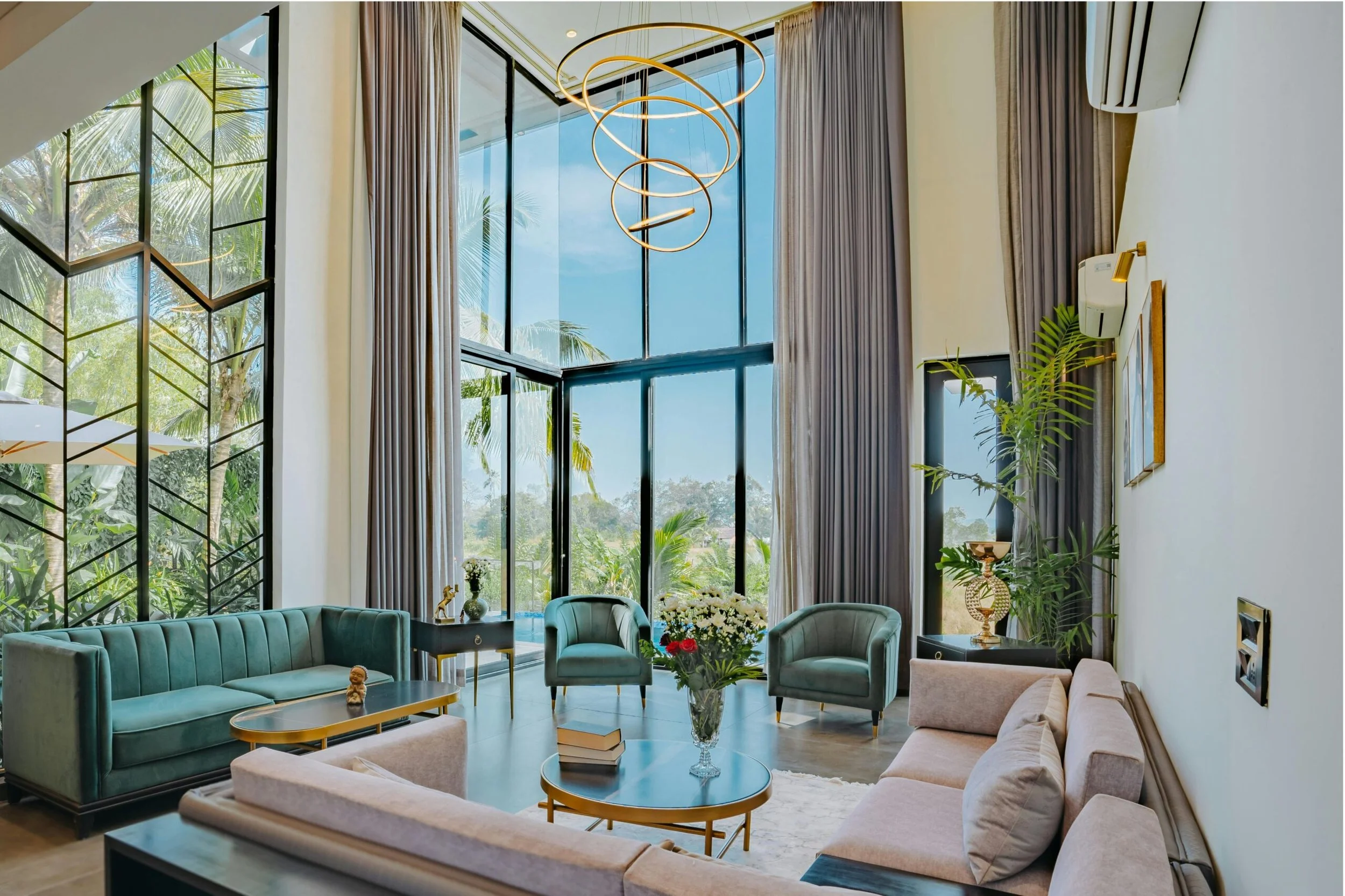 A spacious living room with large floor-to-ceiling windows showing a tropical outdoor scene with palm trees. The room features modern furniture including light blue and beige sofas, a glass coffee table with books, and a floral arrangement, with a St