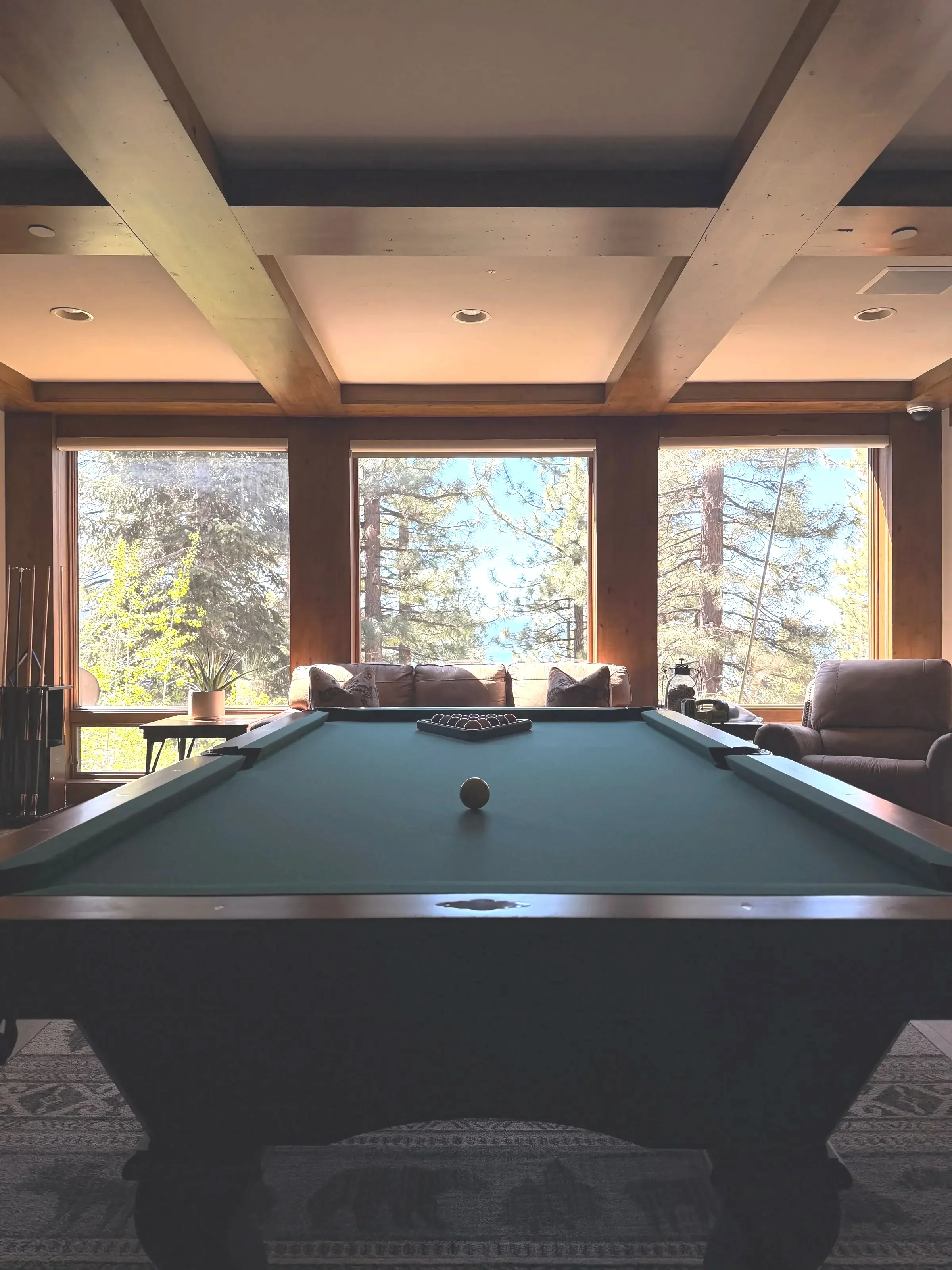 An indoor room featuring a pool table with a white cue ball, positioned centrally. Large windows in the background reveal a view of tall trees and a bright sky. To the right, there is a sofa with cushions and a small side table, establishing a cozy and relaxed atmosphere.