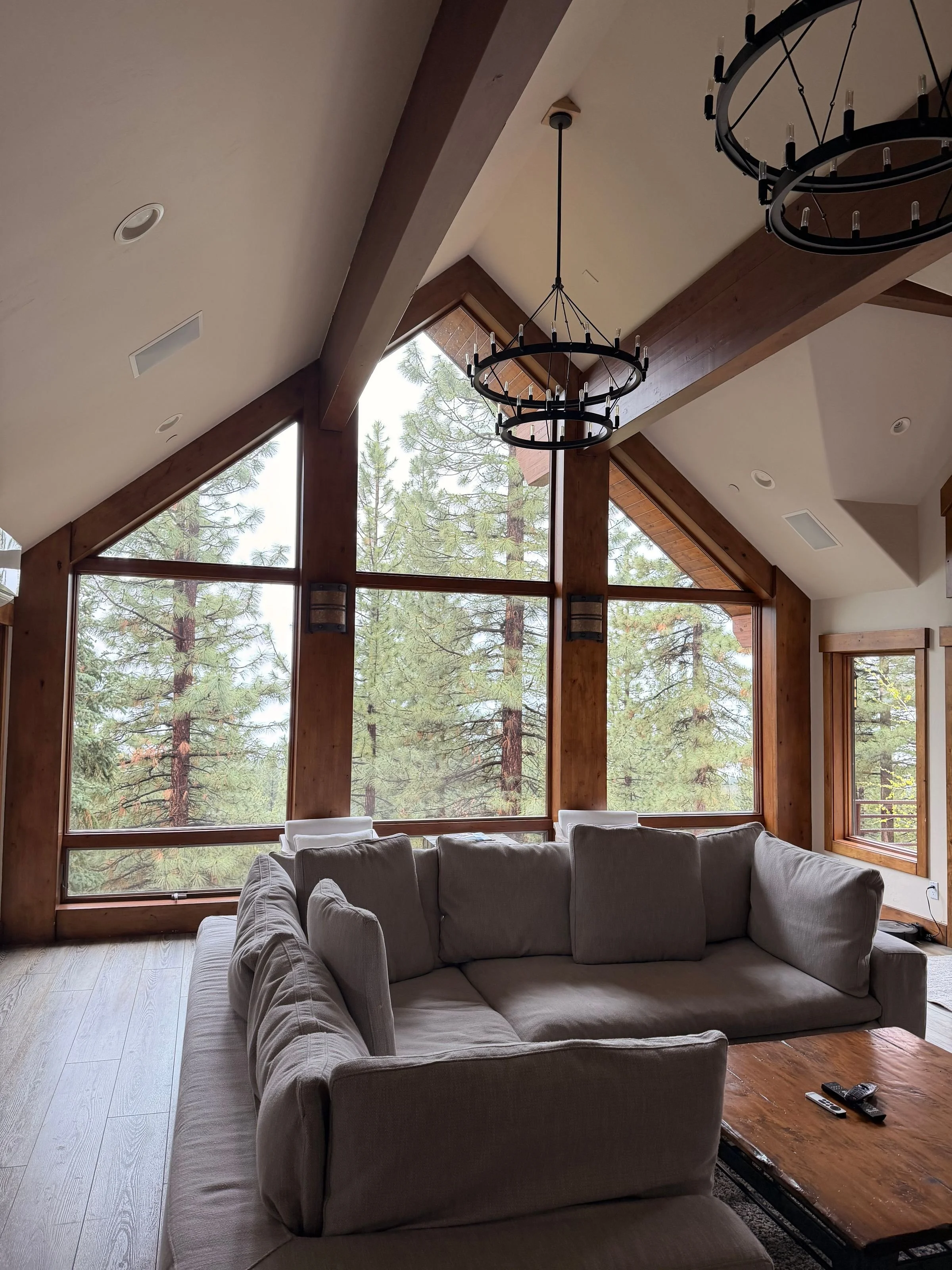 Living room with large windows overlooking trees, beige sofas, a wooden coffee table with remote controls, and a black circular chandelier hanging from the ceiling.