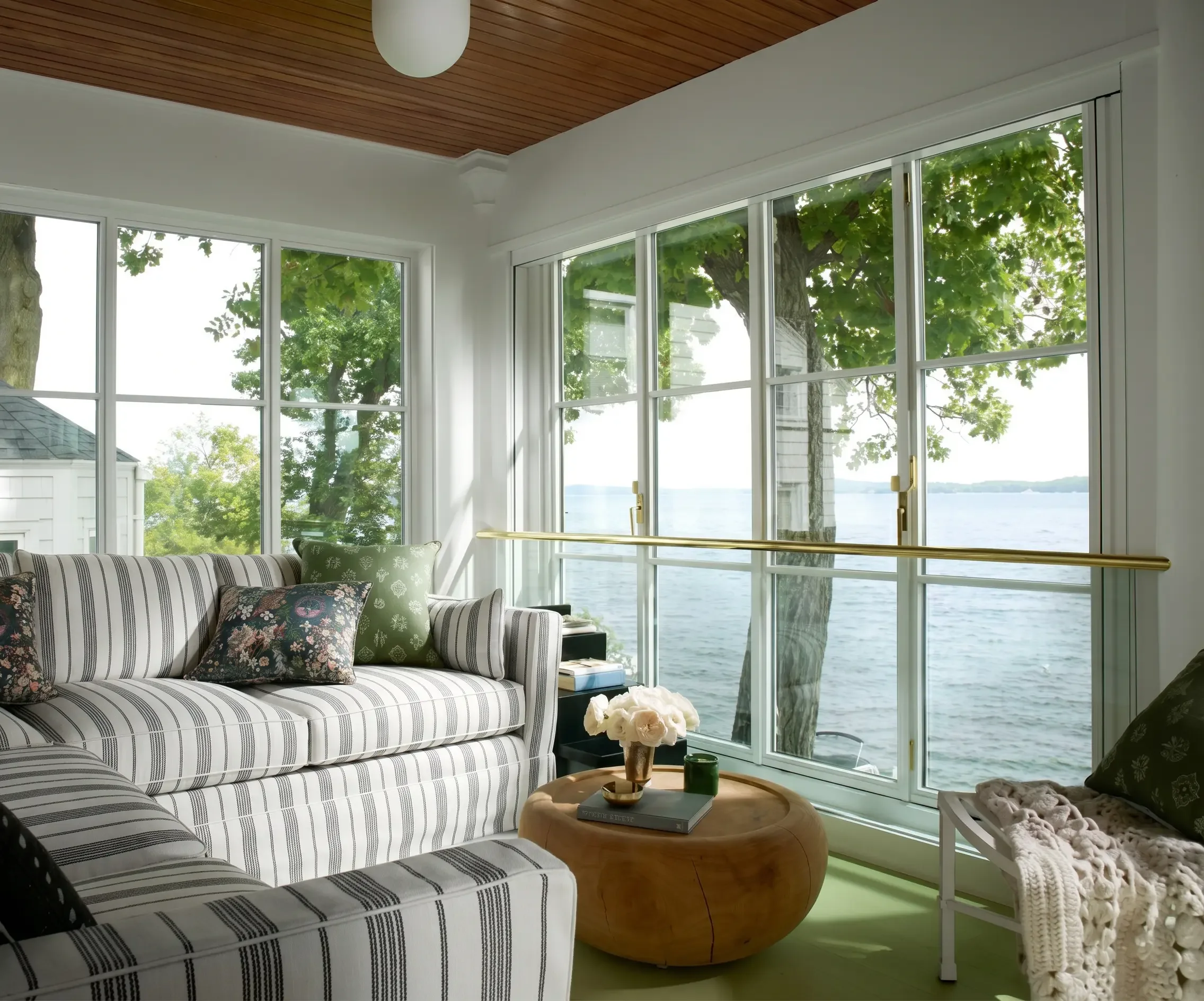 A living room with large windows showing green trees and water outside. The room has a striped sofa with floral cushions, a wooden side table with a flower vase and books, and a small white table with a crochet blanket. The ceiling is wooden.