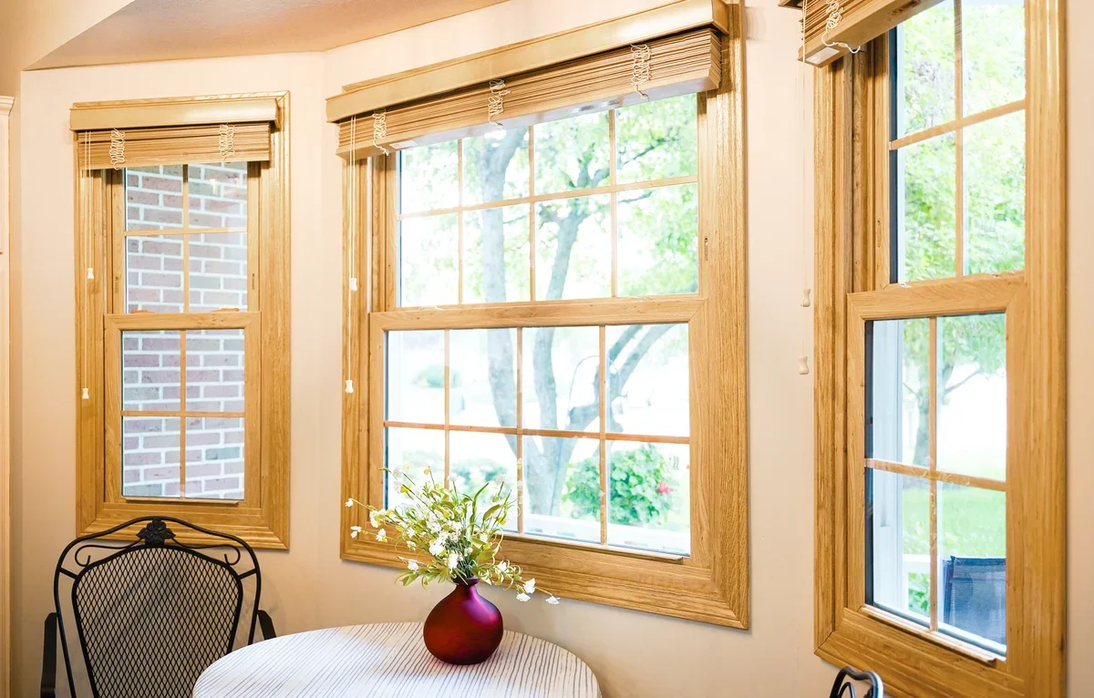 Sunlit room with three large windows with wooden frames and beige blinds, a round table with a red vase of white flowers, and a black chair.