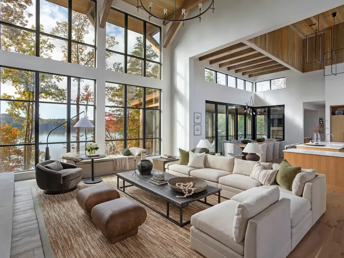 Living room with high glass windows overlooking trees and lake, modern furniture, neutral color scheme, wooden ceiling beams, and decorative accents.