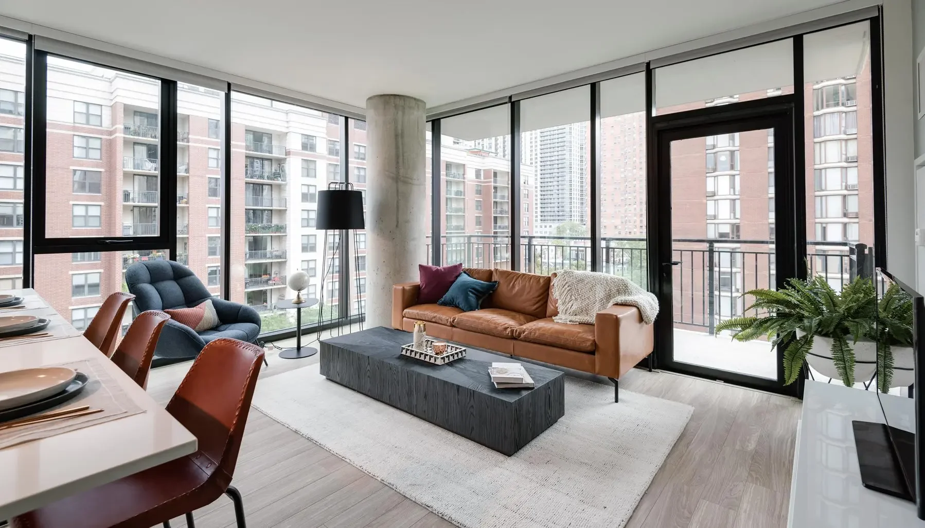Modern living room with a brown leather sofa, a black armchair, and floor-to-ceiling windows with a city view, decorated with pillows, a blanket, a coffee table with items, and potted plants.