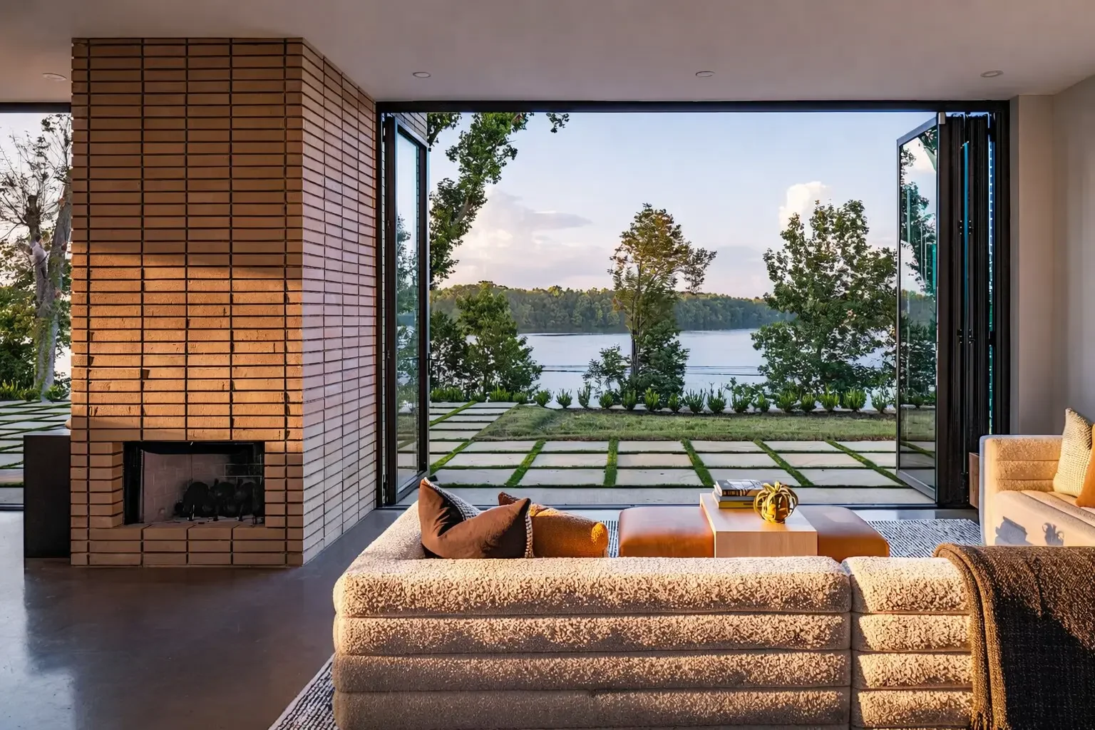 Living room with large open glass doors leading to a lakeside outdoor patio, furnished with a beige textured sofa, pillows, and a wooden coffee table, with a brick fireplace on the left and trees visible through the doors.