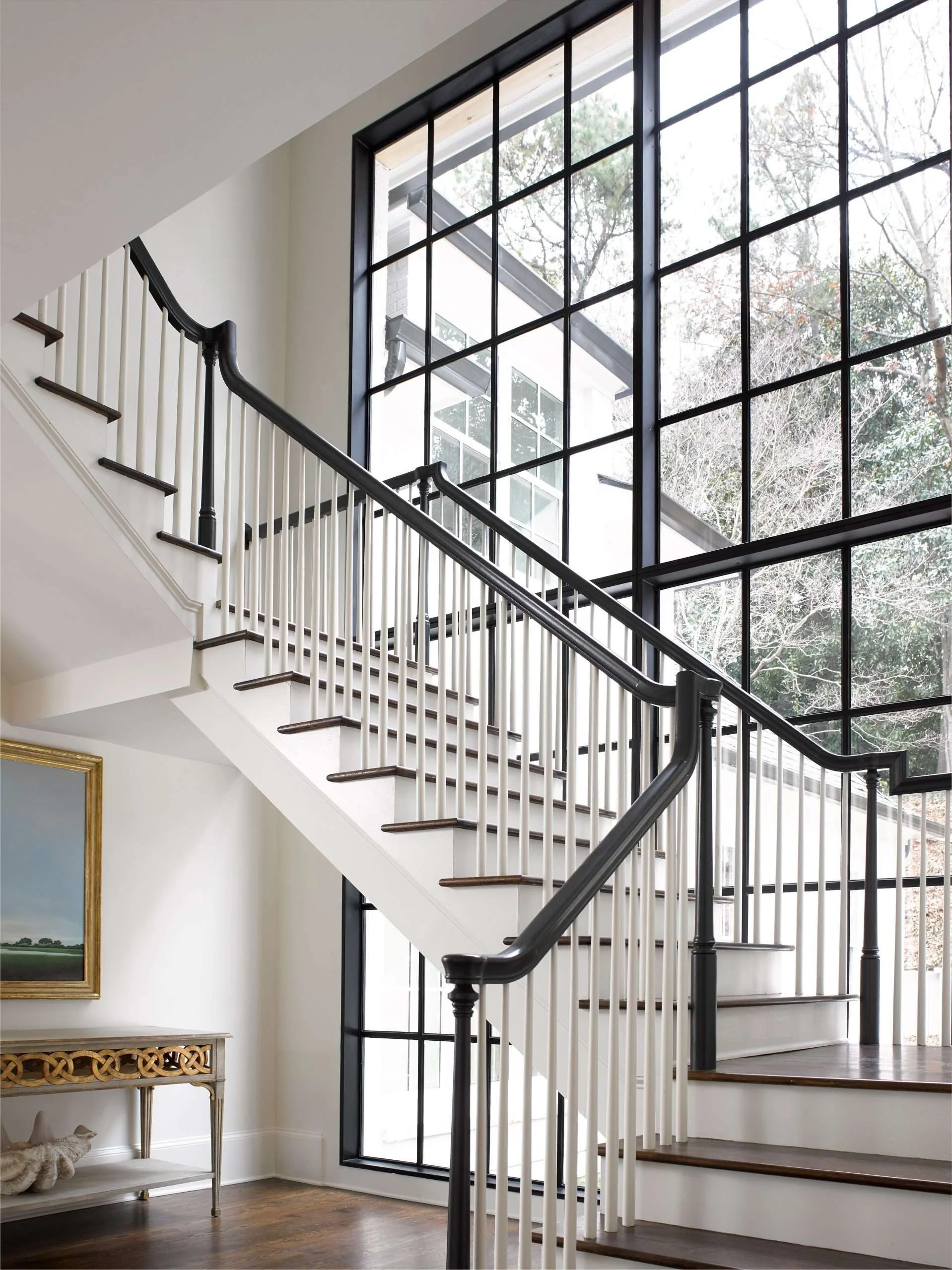Interior view of a staircase with dark wood steps and white railings, next to large black-framed windows showing trees outside, with part of a colorful painting on the wall and a decorative table underneath.