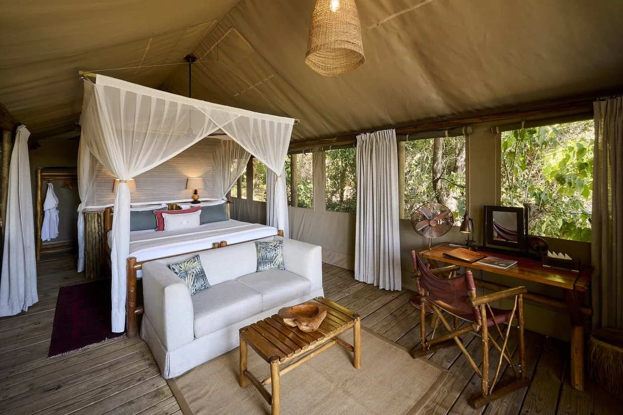A cozy bedroom with a large bed with green and white pillows, a beige sofa with multicolored pillows, wooden floors, windows with a view of trees, a wooden desk with a hat on a chair, and a woven trunk at the foot of the bed, all within a rustic tent-like structure with exposed wooden beams.