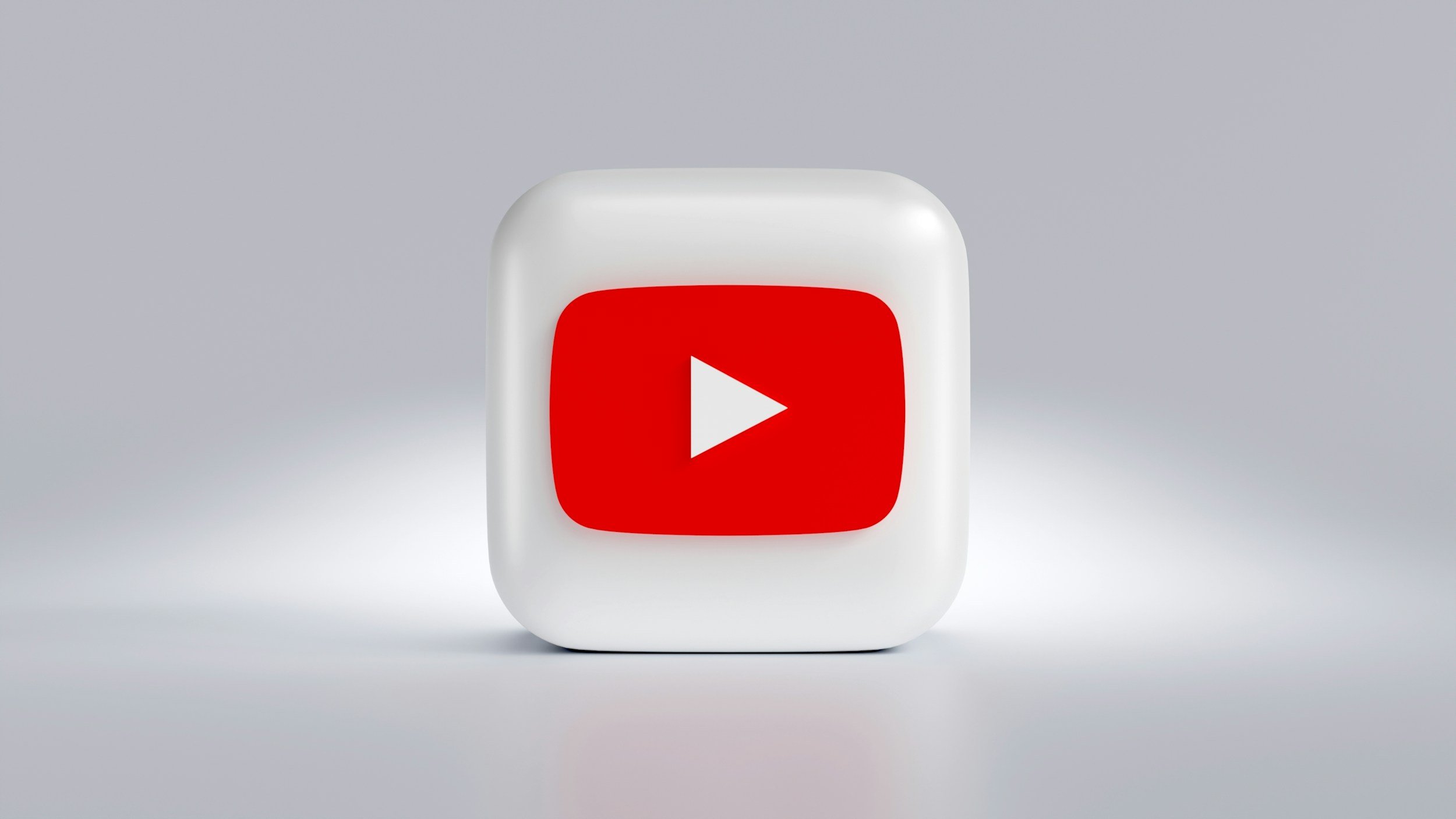 Close-up of a white 3D cube with a rounded edge, featuring the red YouTube play button icon on its front face, set against a light grey background.