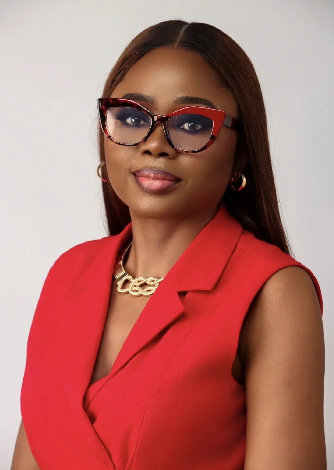 A woman with long brown hair, wearing red and black glasses, a red sleeveless blazer, gold hoop earrings, a gold chain necklace, and makeup that includes lipstick and eye makeup, posing against a plain white background.