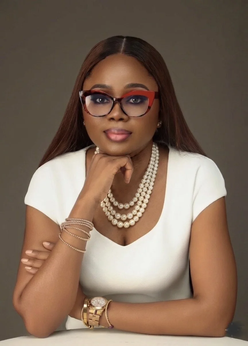Portrait of a woman with long brown hair wearing large red and black glasses, a white dress, layered pearl necklaces, and gold jewelry, sitting with her chin resting on her hand against a neutral background.