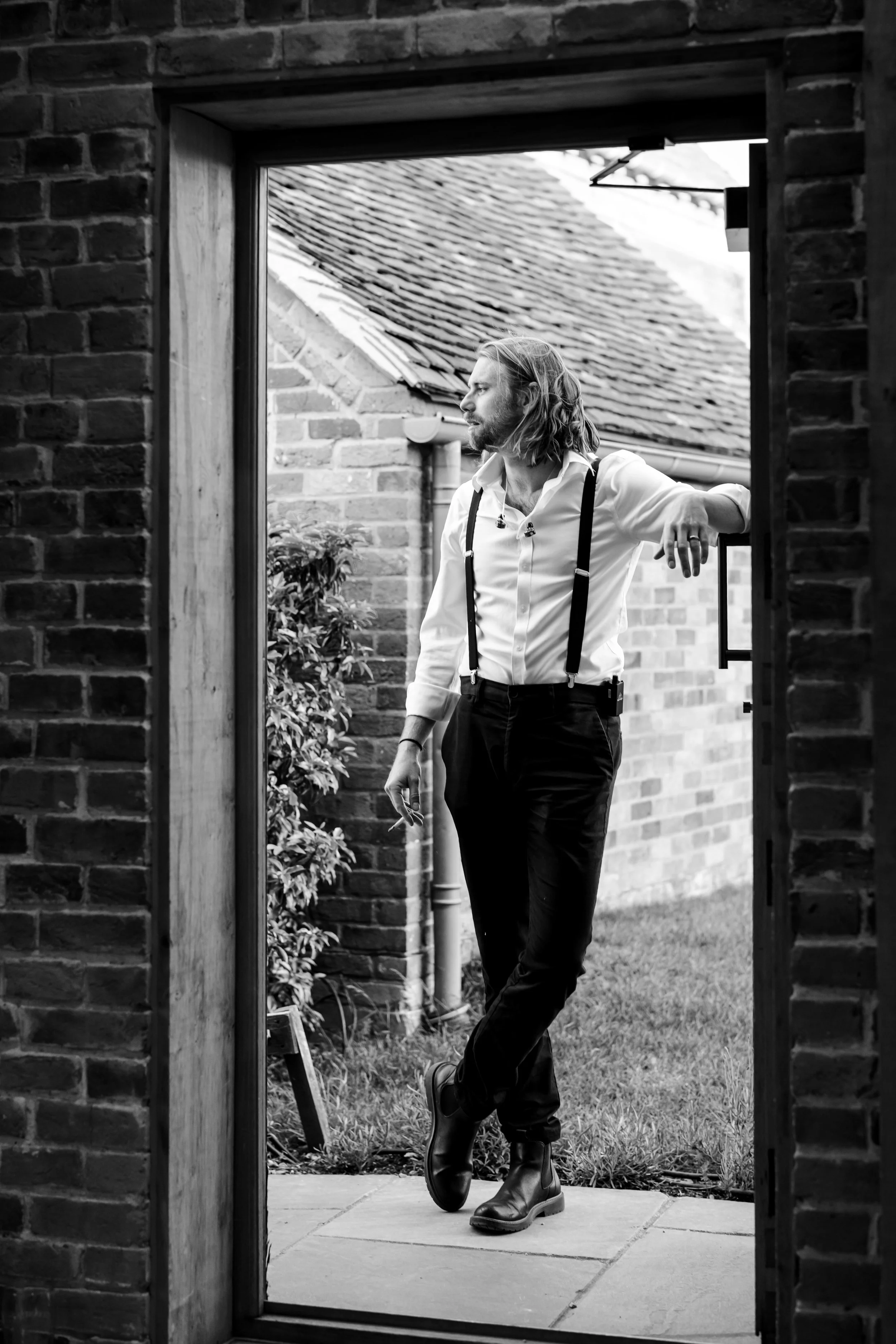 A man with shoulder-length hair and a beard leaning against the doorframe outside, wearing a white shirt, suspenders, black pants, and boots, looking to the side in a brick patio area.